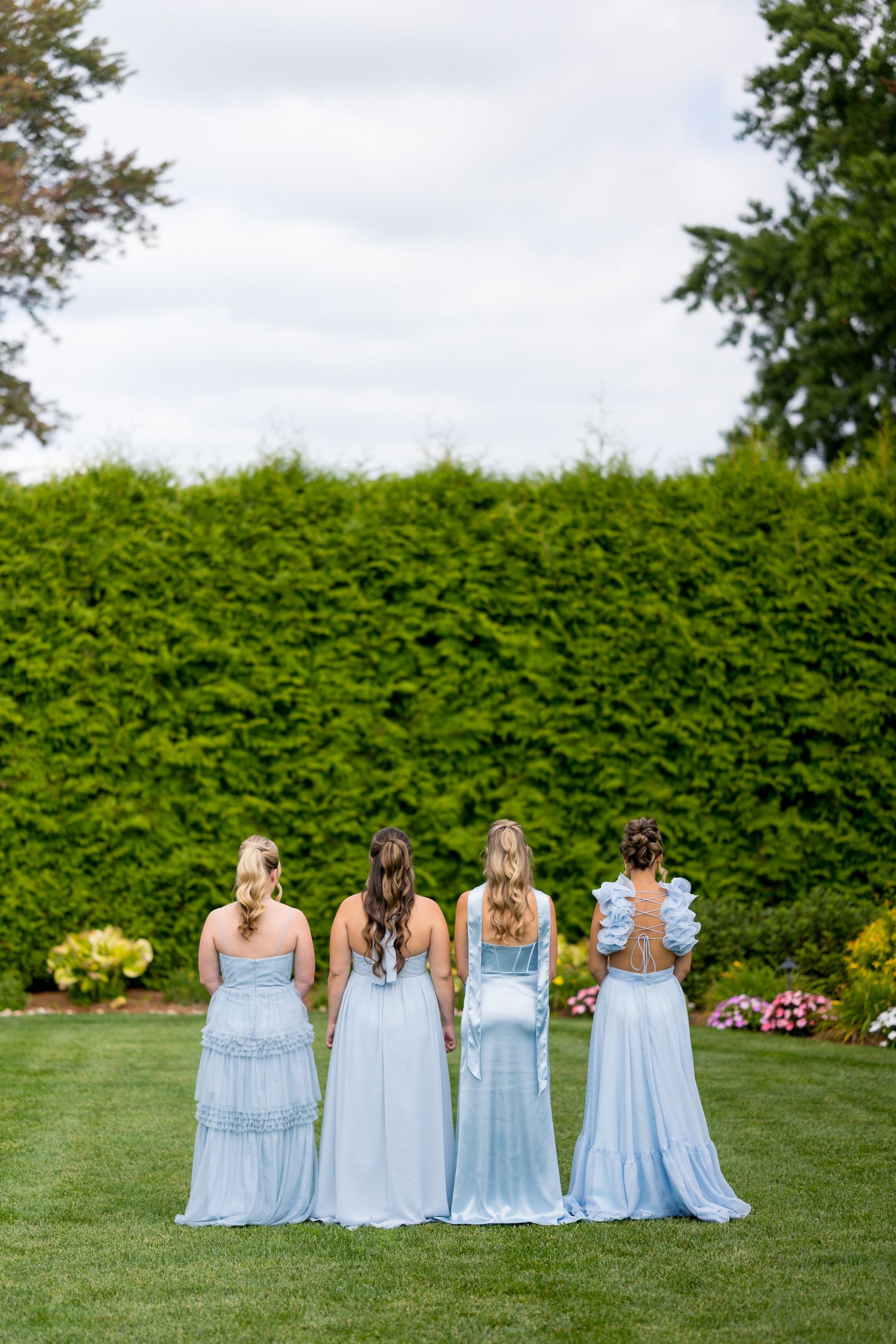 Four bridesmaids in light blue dresses stand with their backs to camera on a manicured lawn with green hedge backdrop.