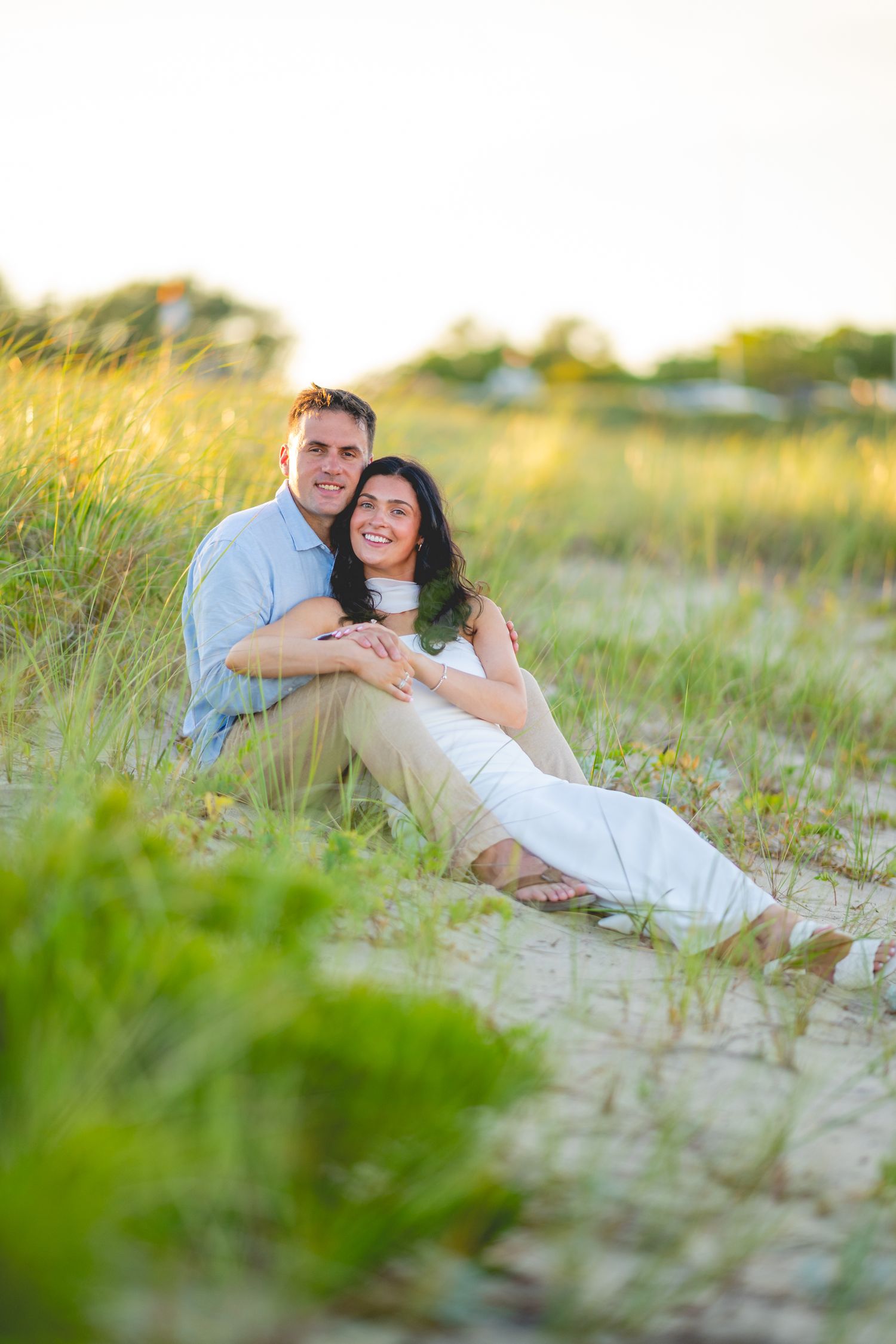 A couple relaxes together on a sandy beach during golden hour with tall grass in the foreground.
