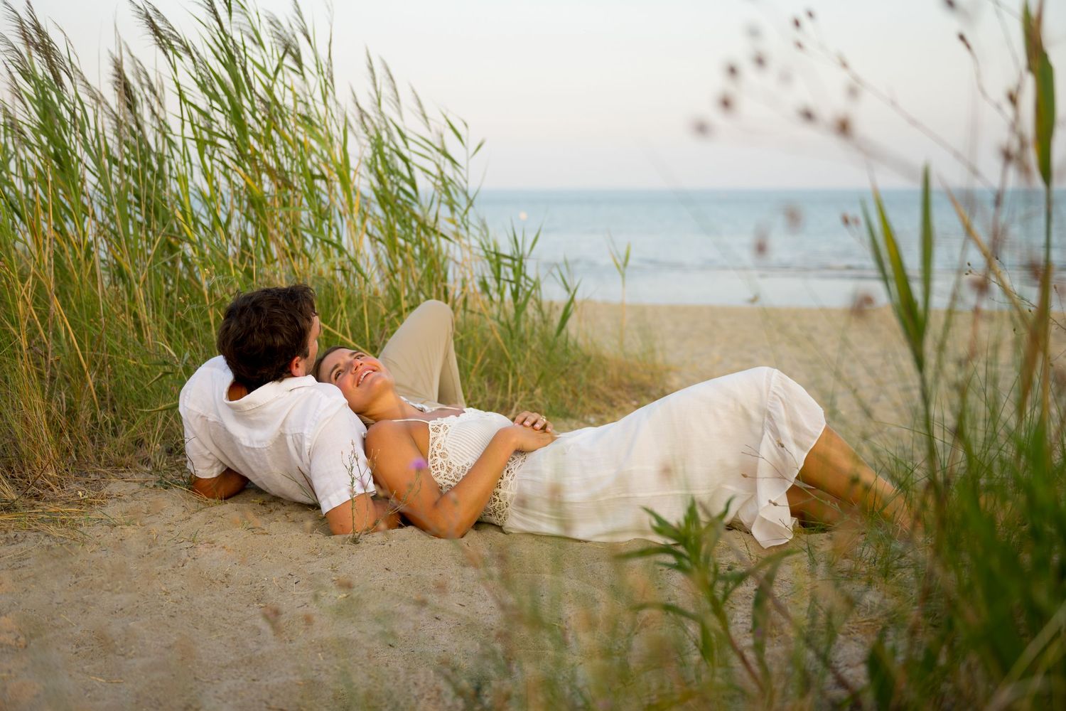A couple relaxes on the sandy beach at sunset among tall sea grass with ocean waves in the background.