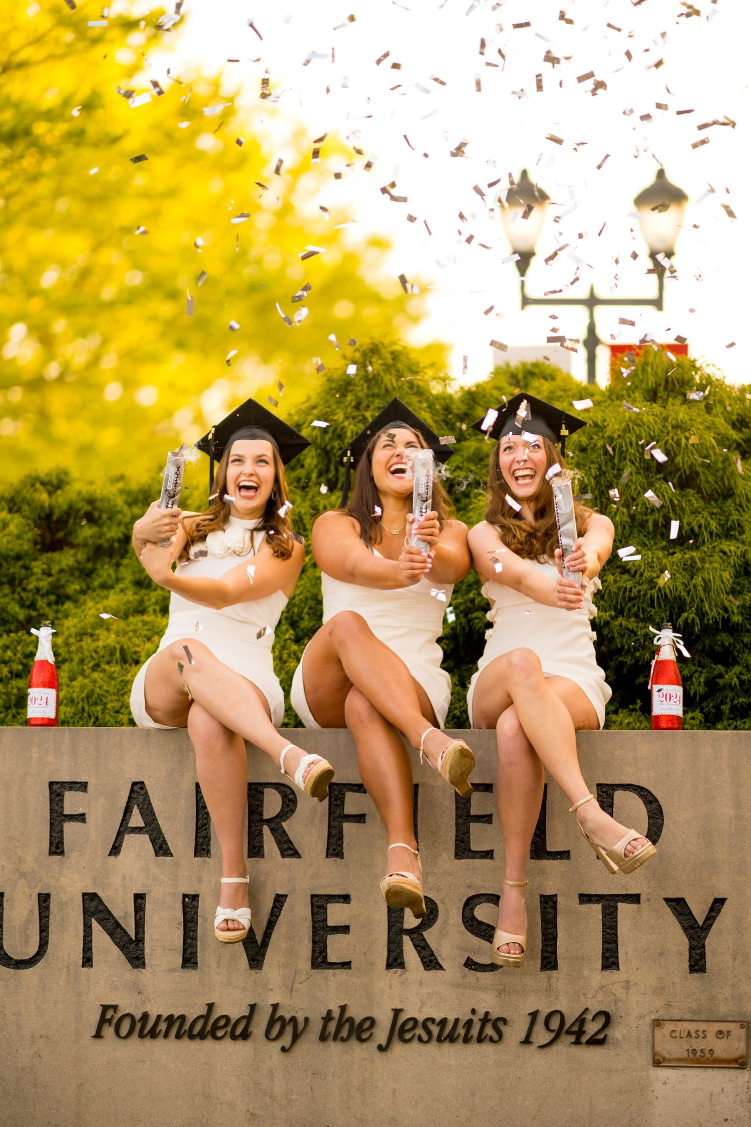 Graduates sitting on Fairfield University sign throwing confetti during celebration photo.