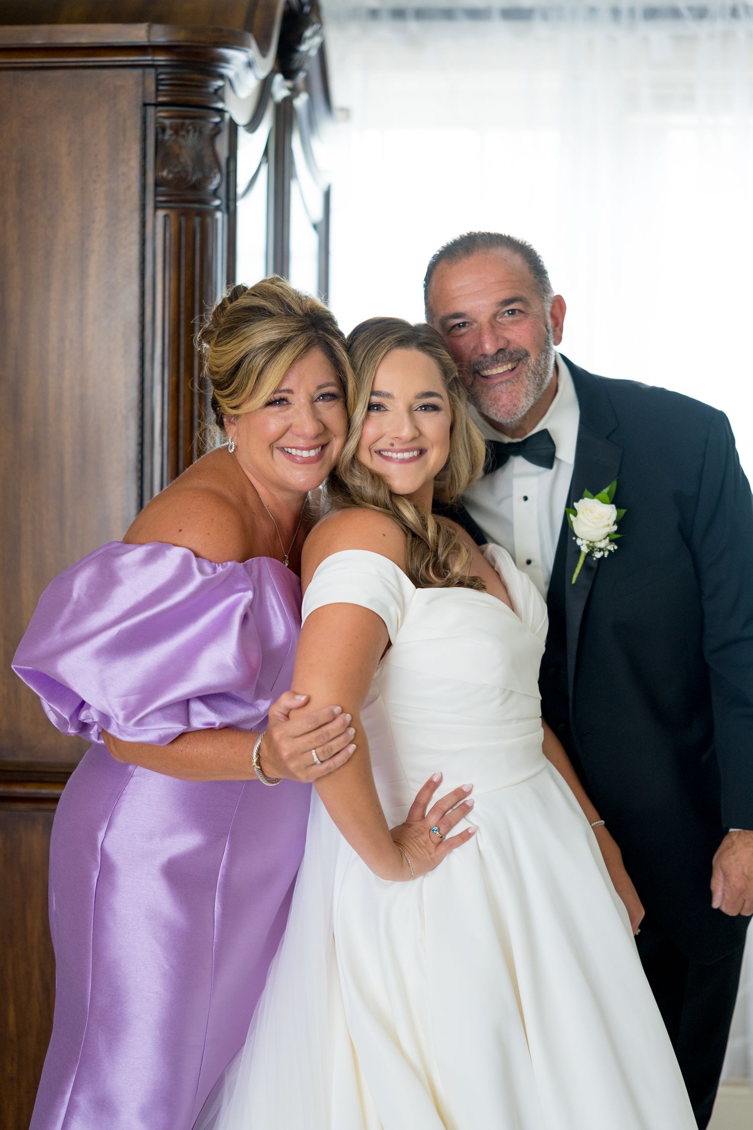 Wedding party members share joyful embraces in formal attire including white and purple dresses.