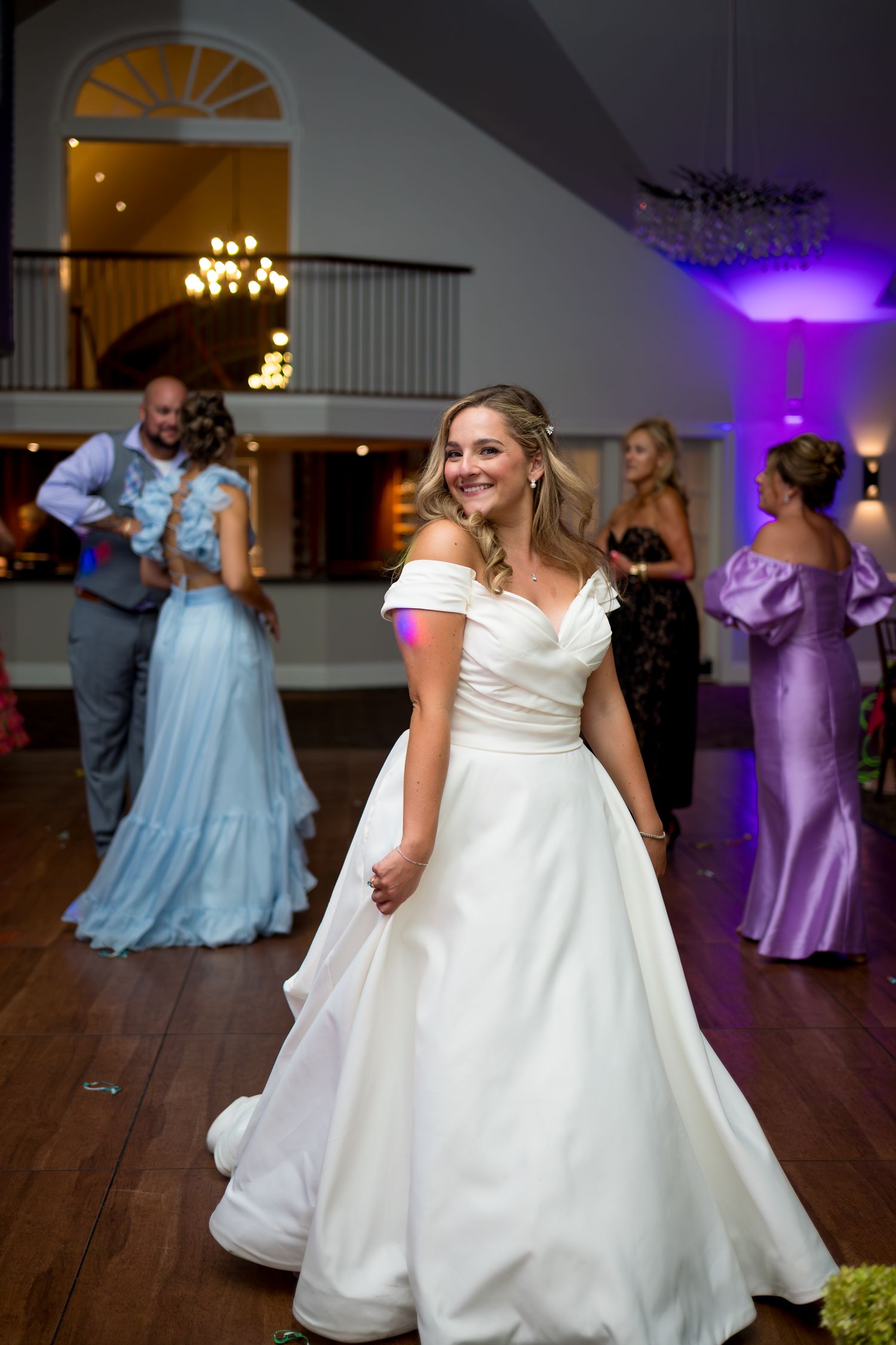 A wedding reception dance floor with guests in pastel blue and purple dresses dancing under chandelier lighting.