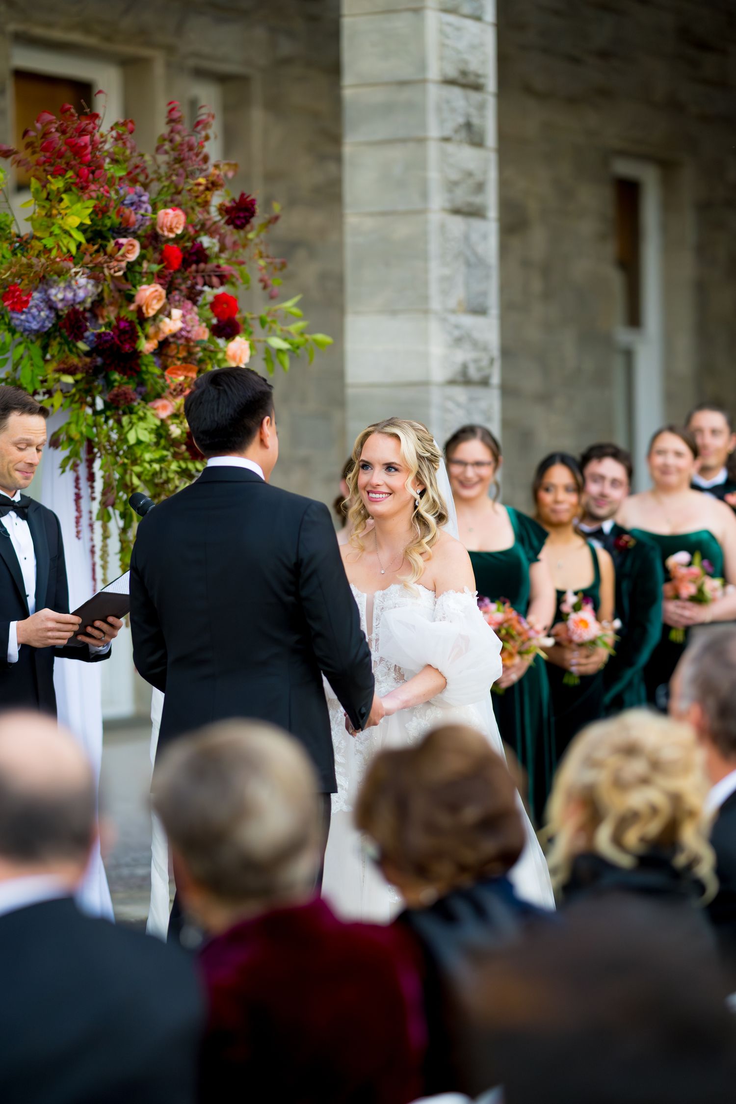 Wedding guests gather around a stone colonnade with vibrant floral arrangements during an elegant outdoor ceremony.