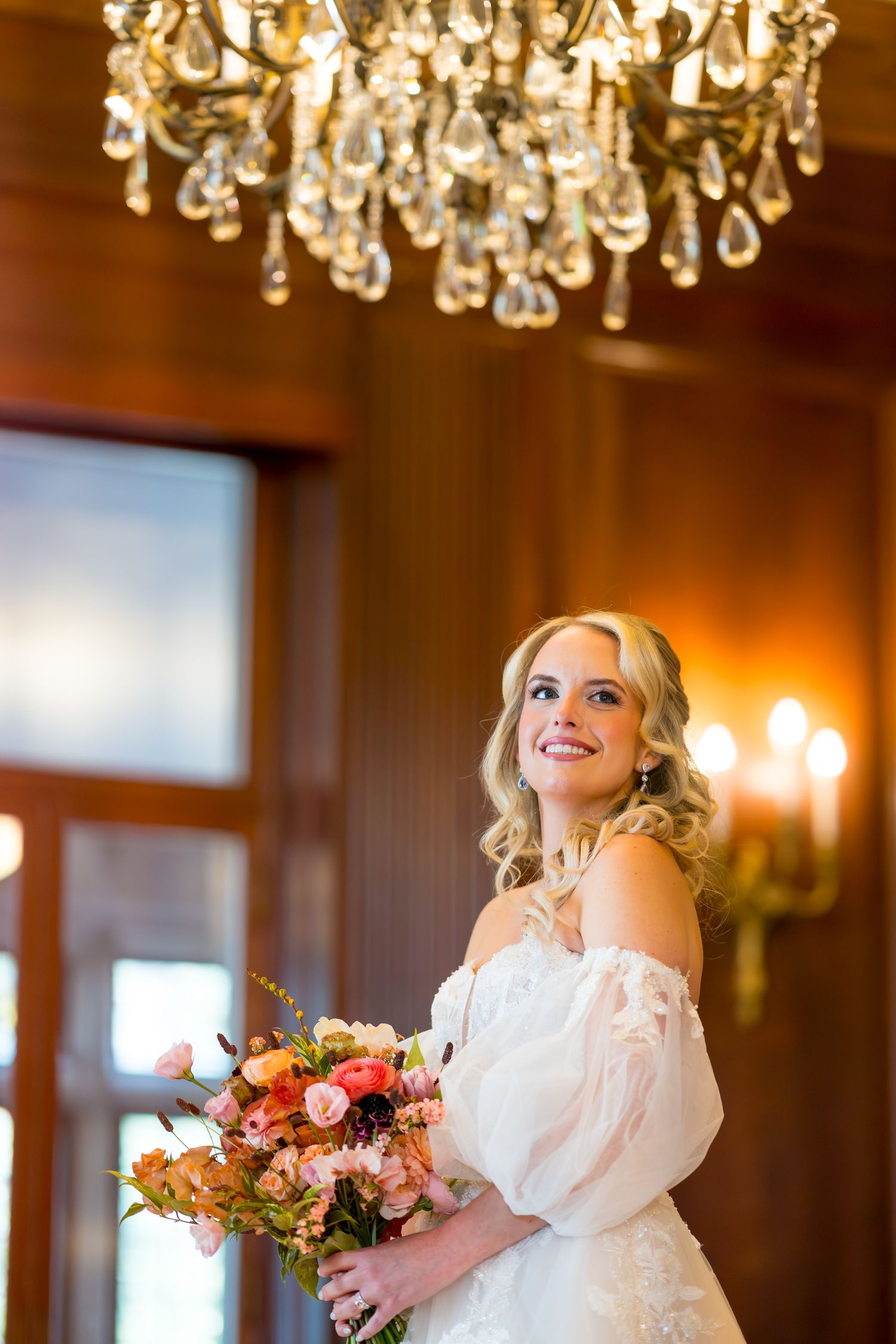 A bride in a white wedding gown holds a colorful bouquet under an elegant crystal chandelier.