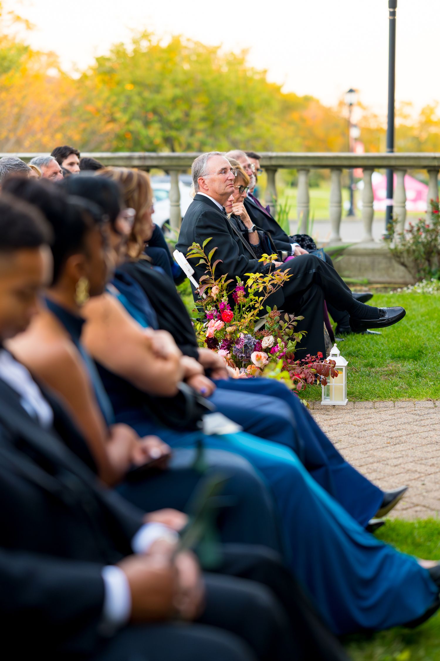 Wedding guests seated in formal attire at an outdoor ceremony with colorful flowers in the background.