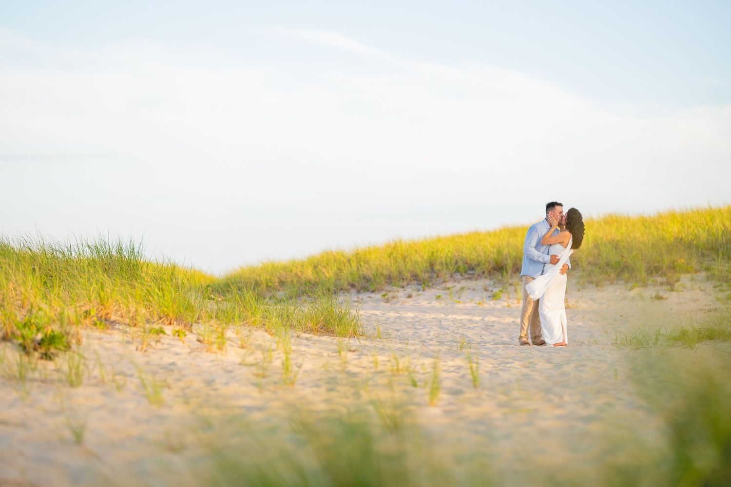 Two figures embrace on a sandy beach path surrounded by golden wildflowers and dunes under soft sunlight.