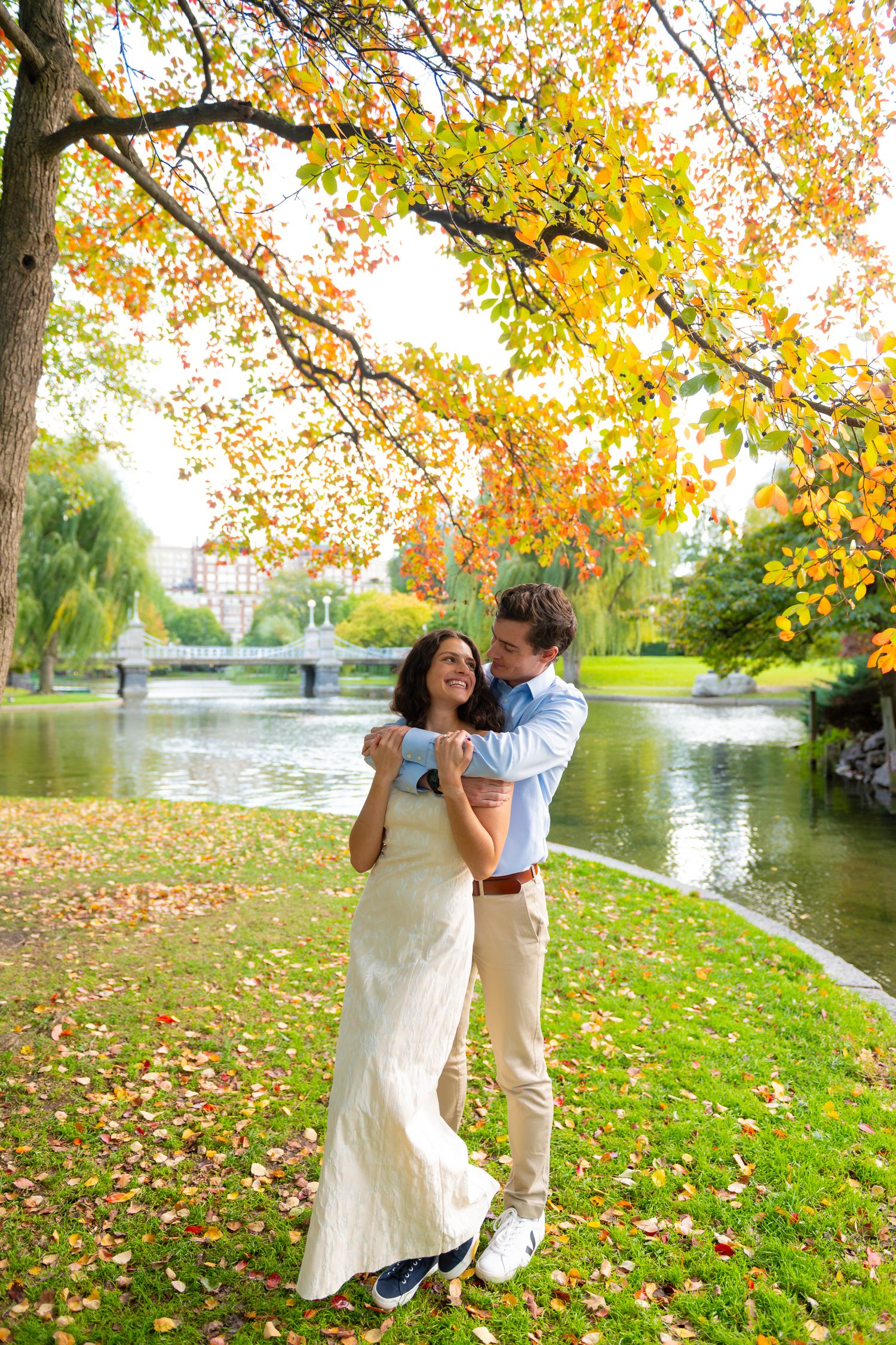 A couple dances together on grass near a lake with autumn foliage in the background at Boston Public Gardens.