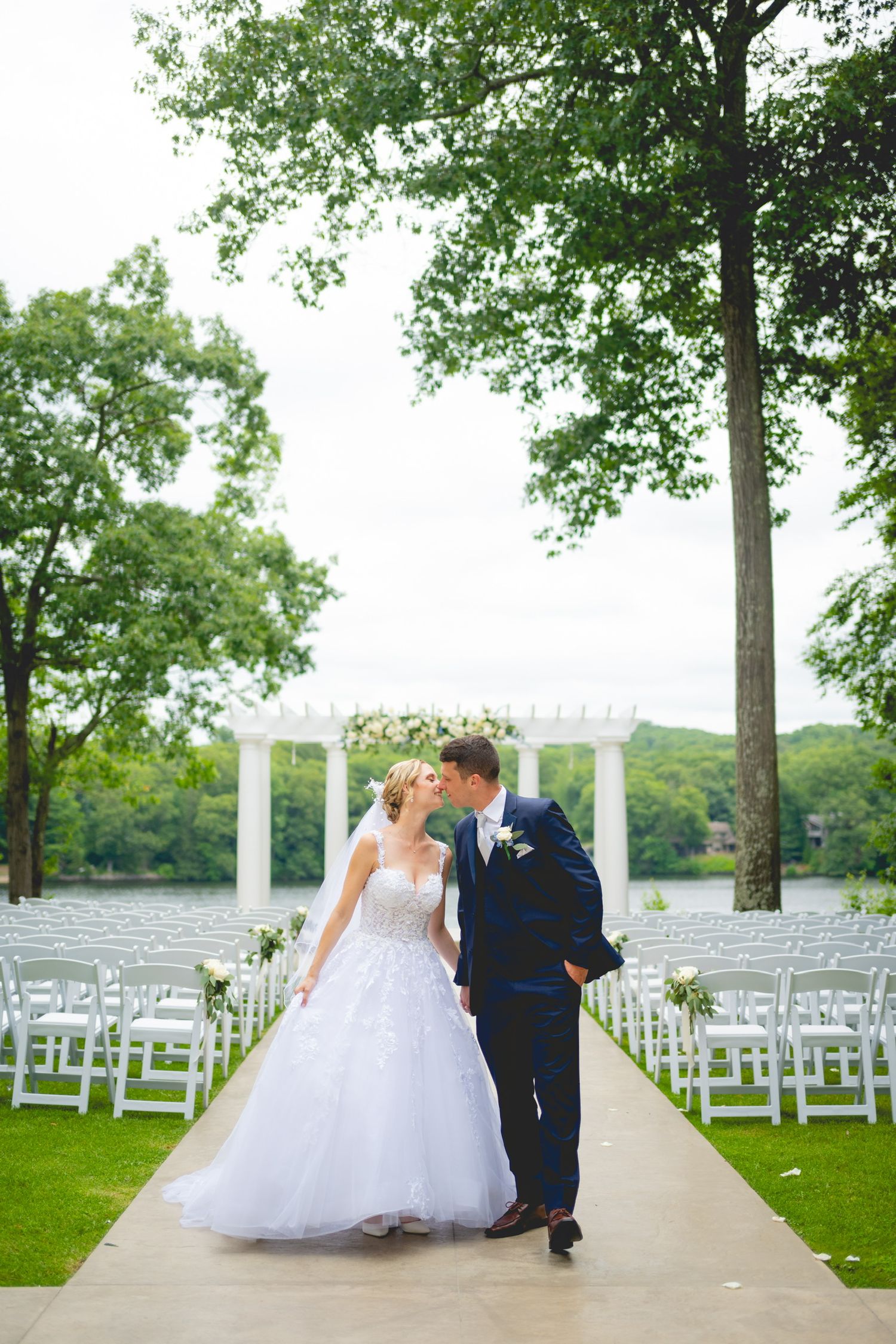 Wedding couple walks along a tree-lined outdoor ceremony setup with white chairs.