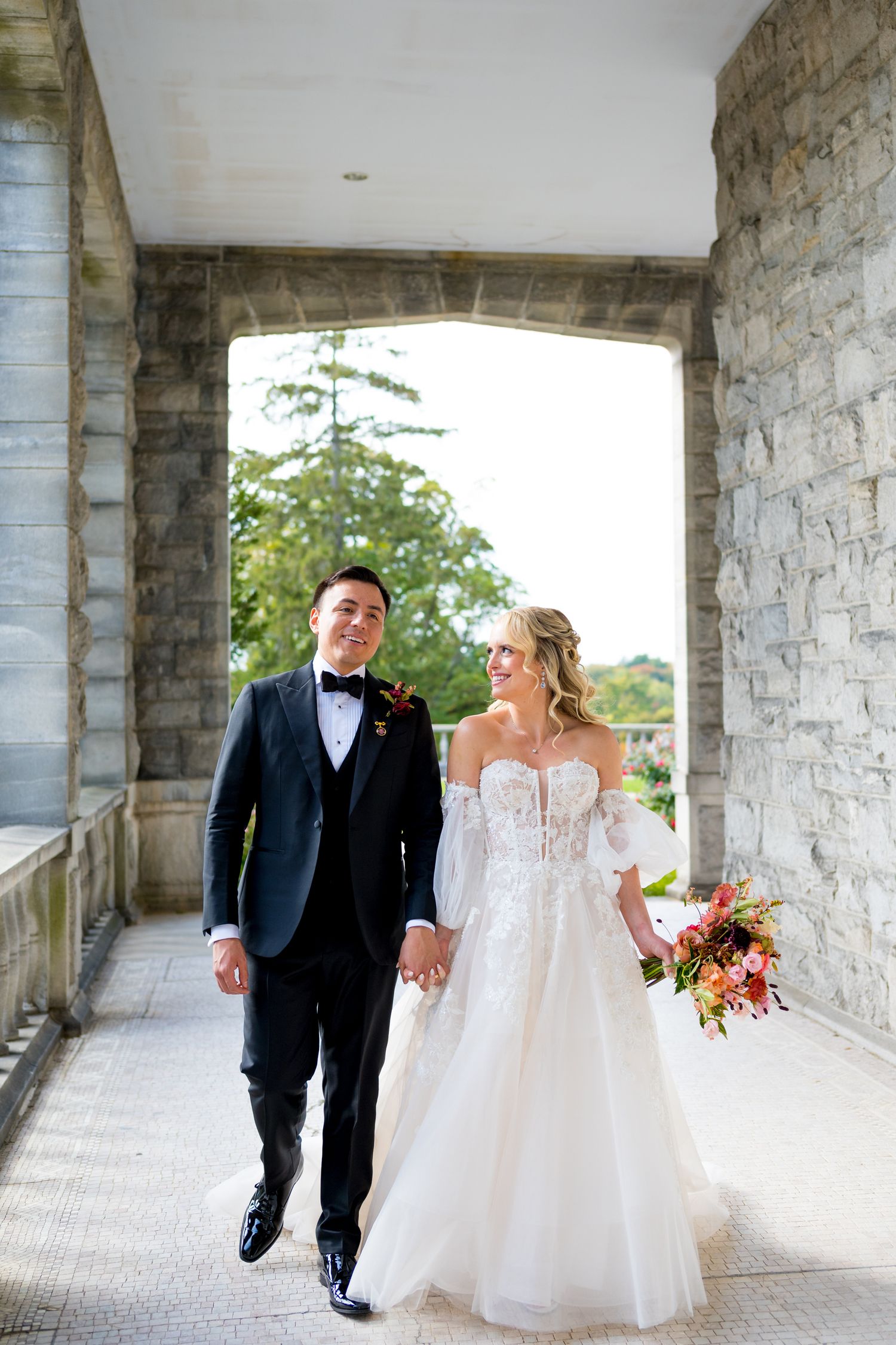 Wedding couple walking and laughing under stone archway colonnade.