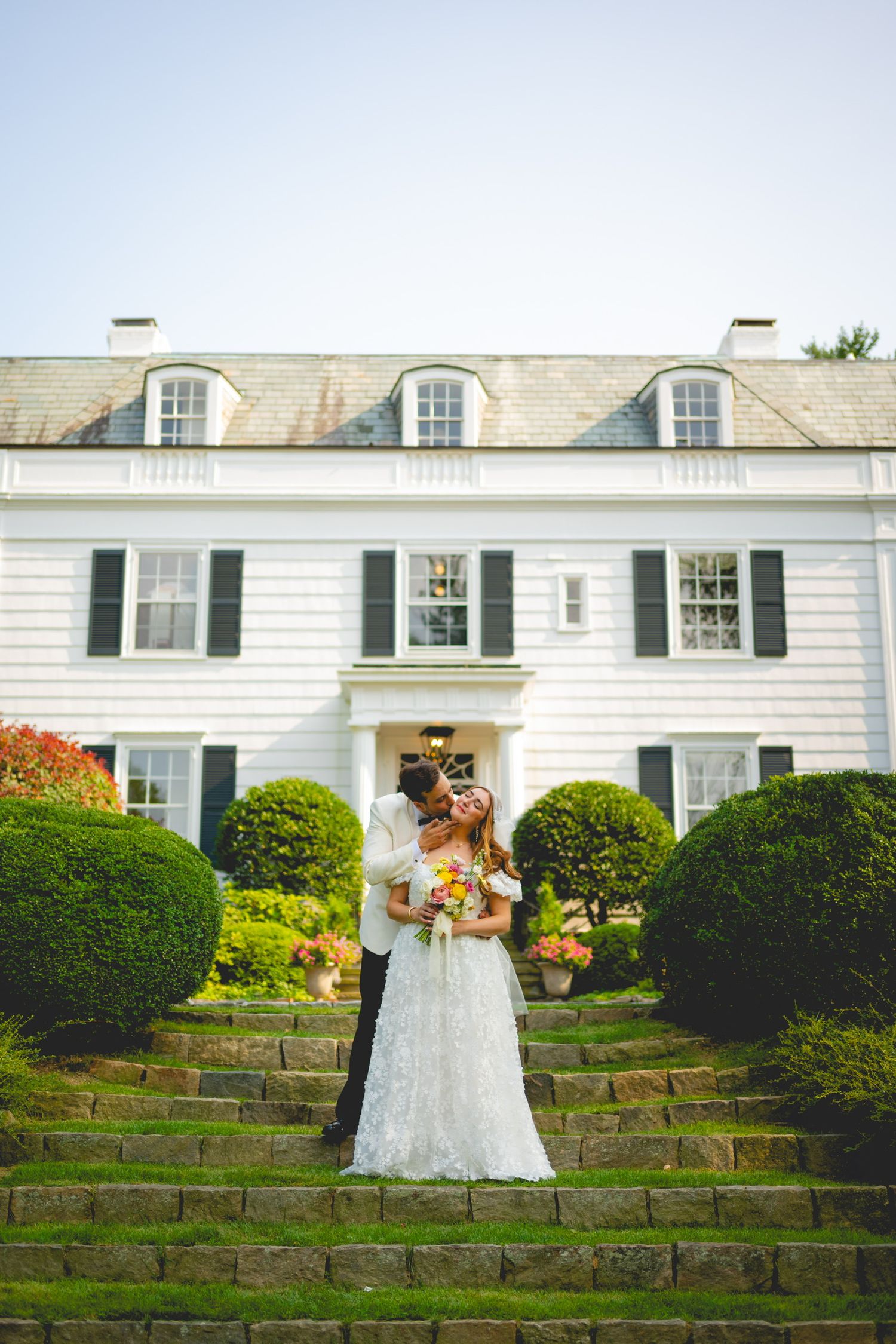 Wedding couple poses on stone steps in front of elegant white colonial-style building.