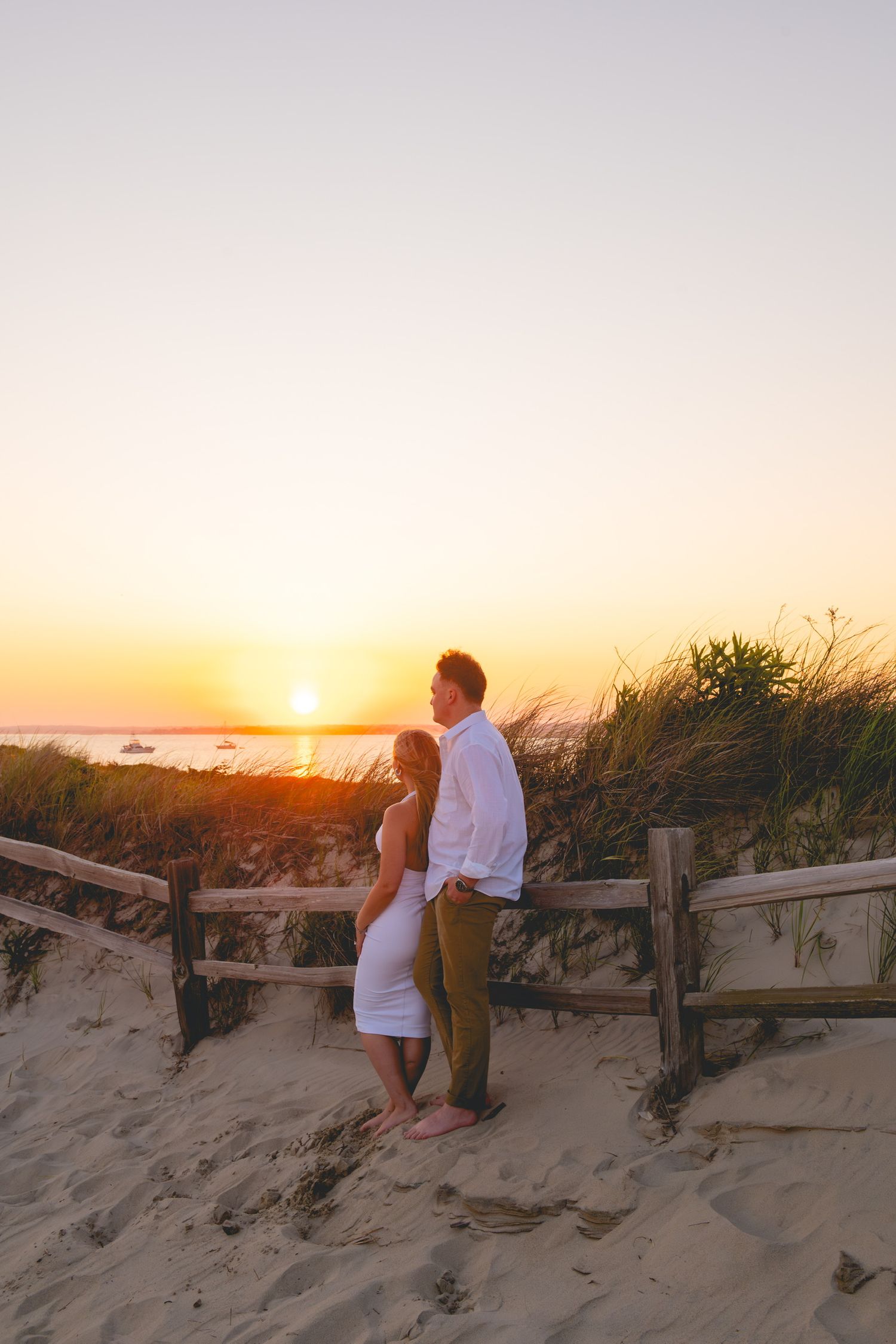 A couple watches the sunset together on sandy dunes with wooden fencing in Watch Hill, Rhode Island.