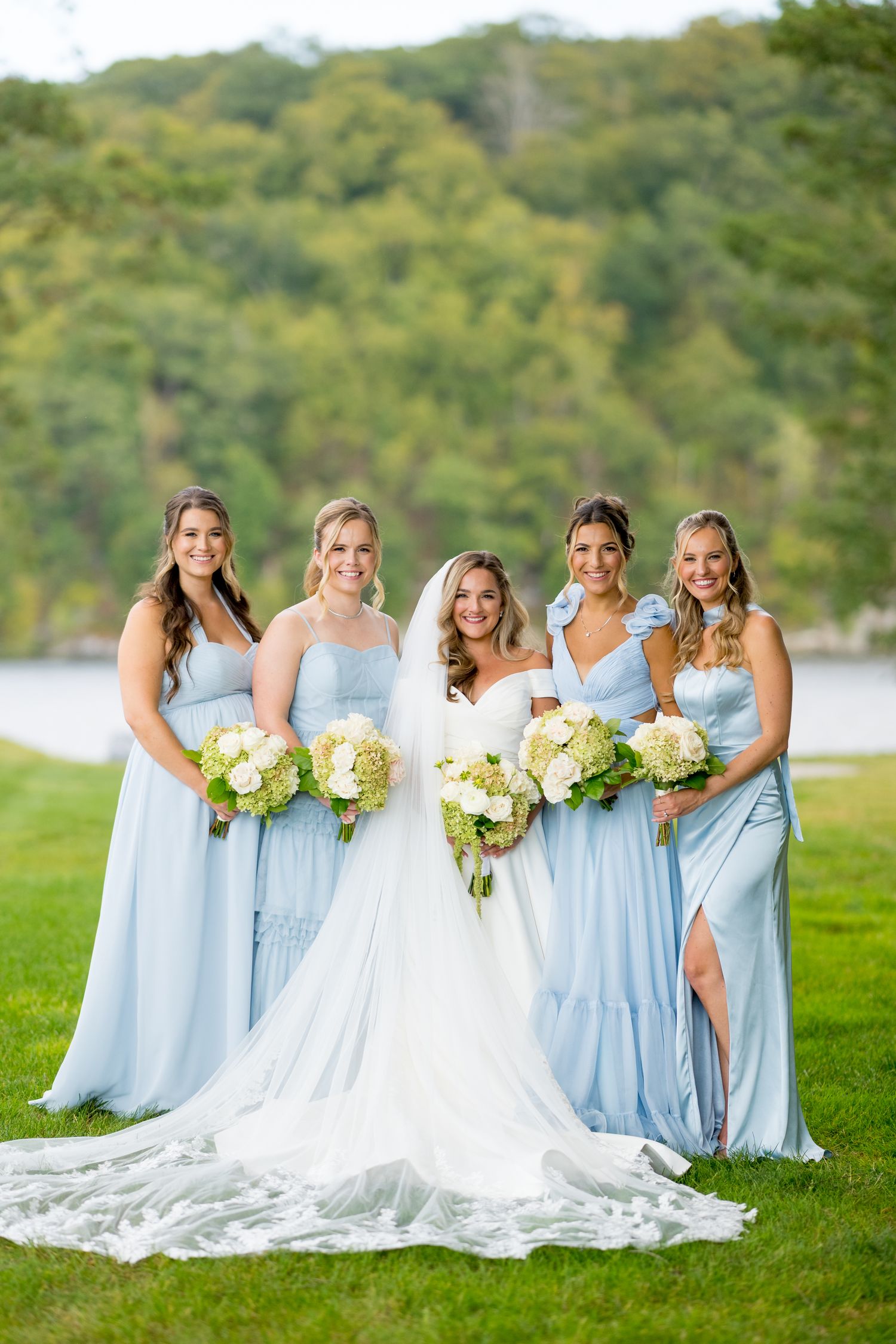 Bridesmaids in light blue dresses stand together with the bride in a white gown on a grassy lawn by a lake.