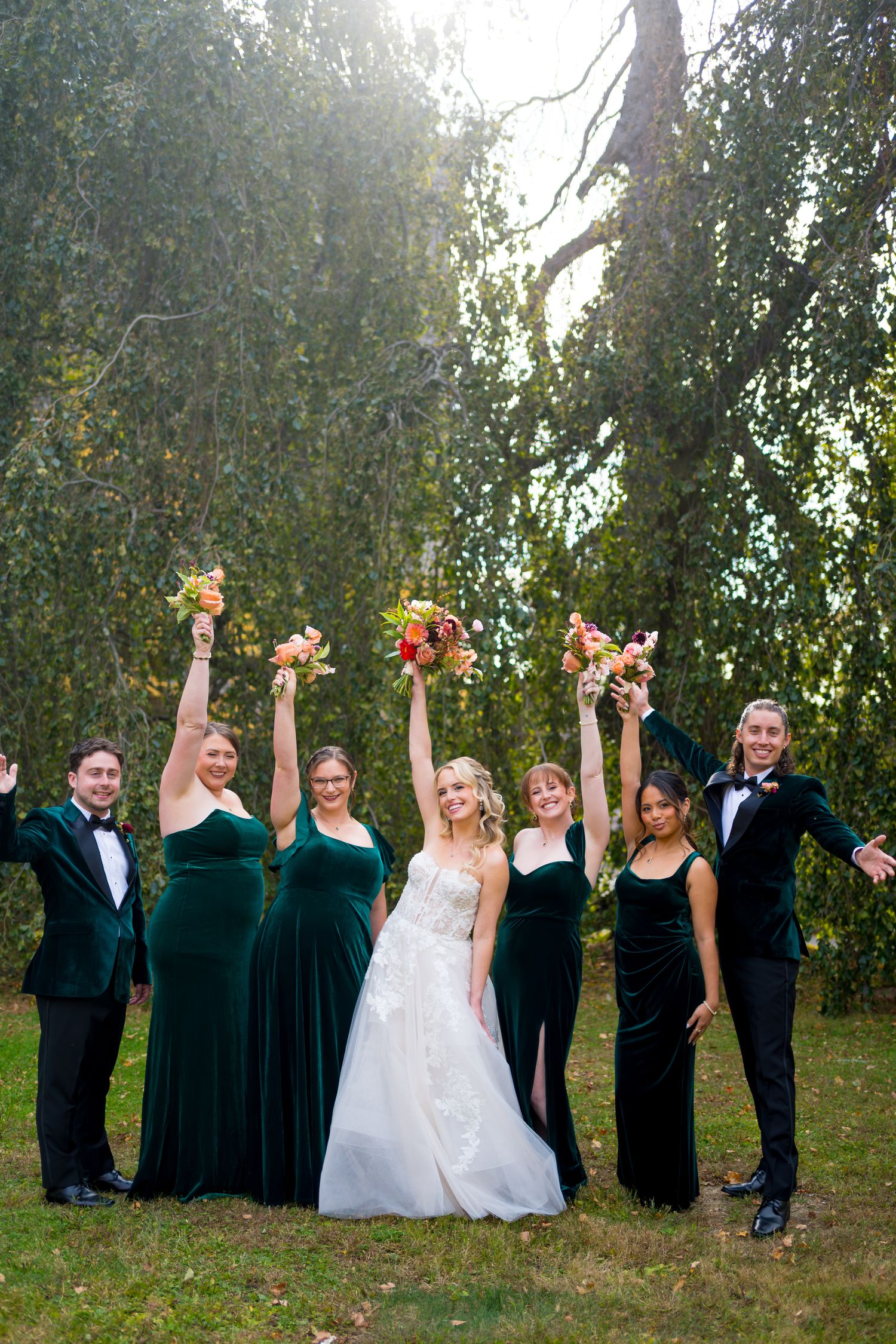 Wedding party in formal attire poses playfully with raised arms in an outdoor garden setting at sunset.