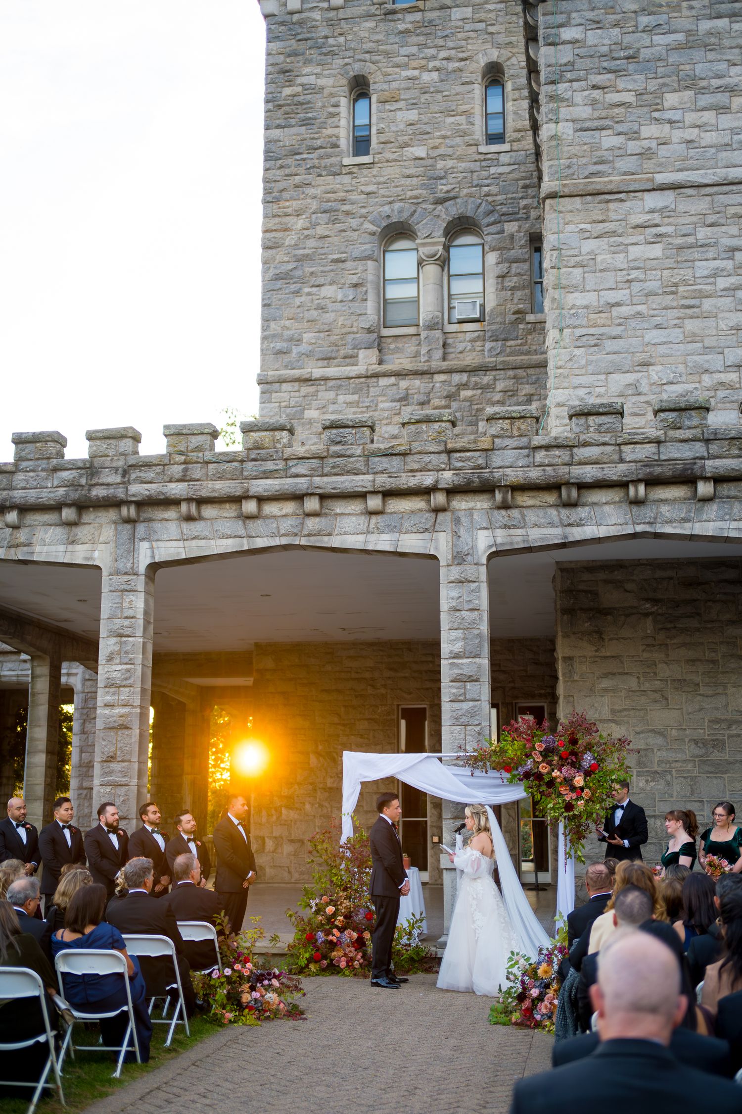 Wedding ceremony taking place at sunset under stone archways of a castle-like venue with guests seated on white chairs.