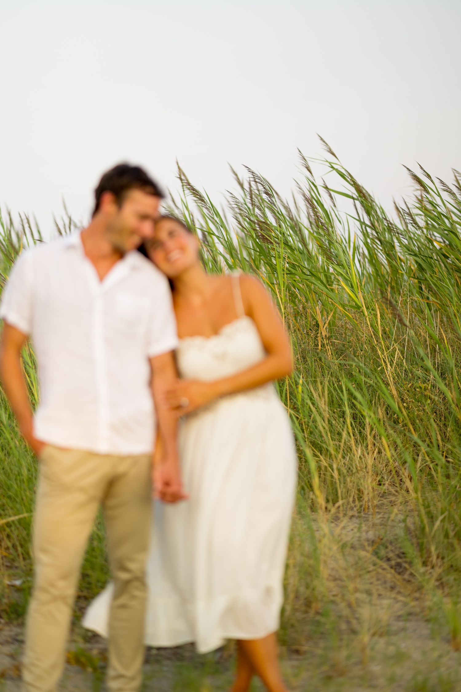 Couple in white and khaki outfits share romantic moment among tall beach grass.