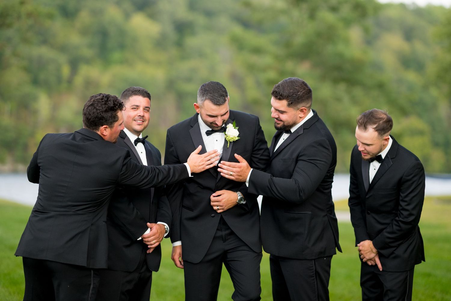 Groomsmen in black tuxedos sharing laughs and embraces outdoors.
