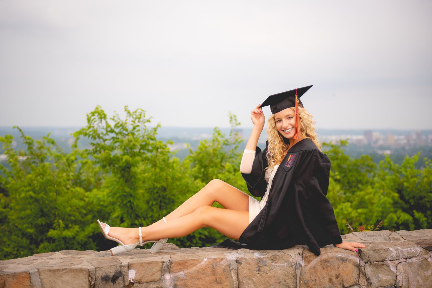 Graduate in black cap and gown sits casually on stone wall overlooking scenic view for graduation photos.