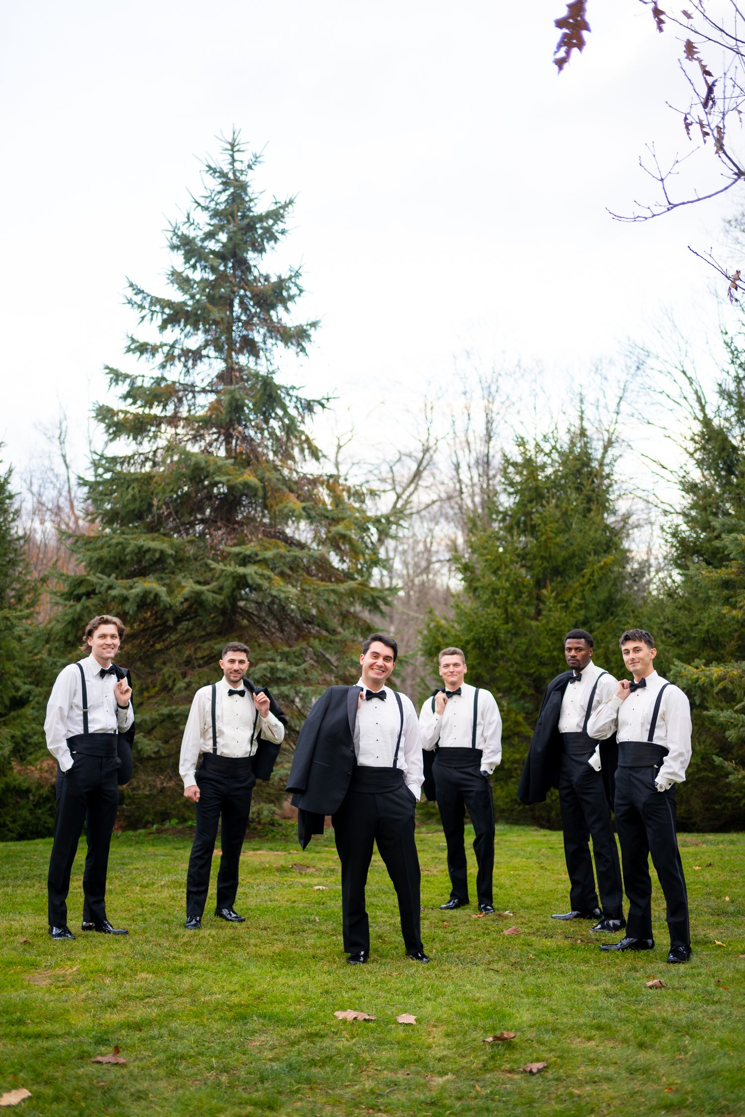 Groomsmen in suspenders and bowties pose casually on a grassy hill for wedding party photos.
