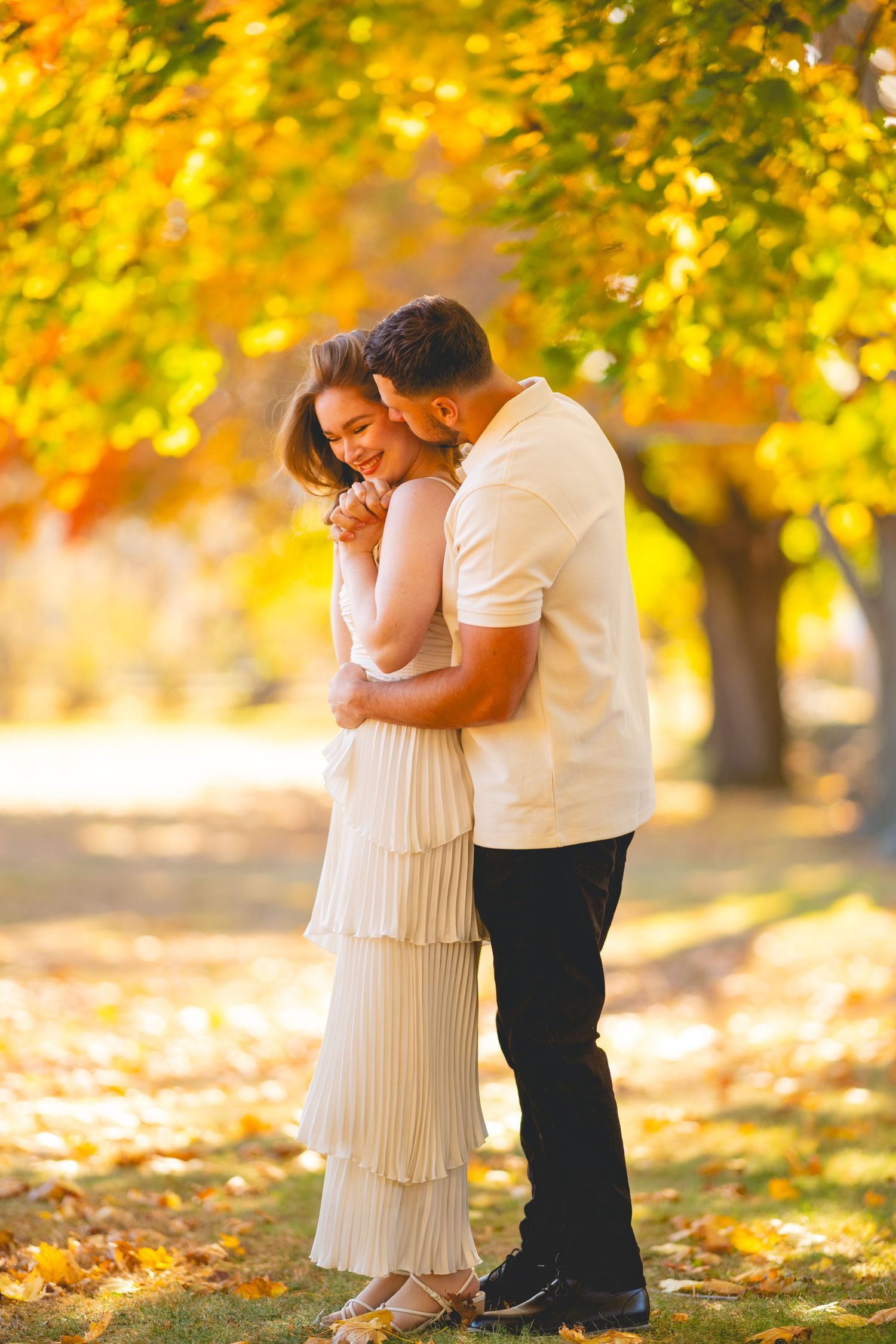 A romantic couple embraces under golden autumn trees with sunlight filtering through the leaves.
