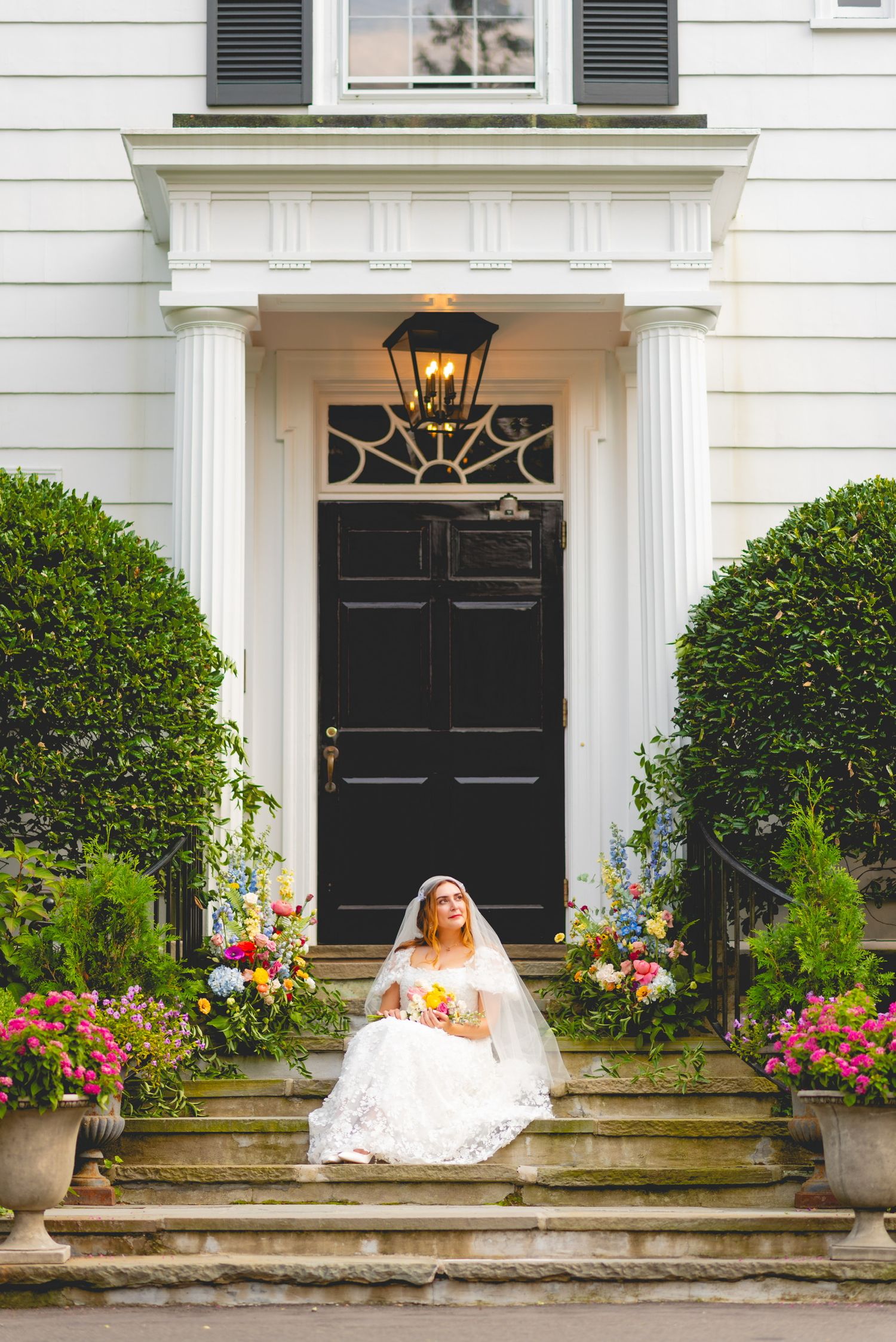 Wedding portrait on white colonial house steps with black door and colorful flower gardens.