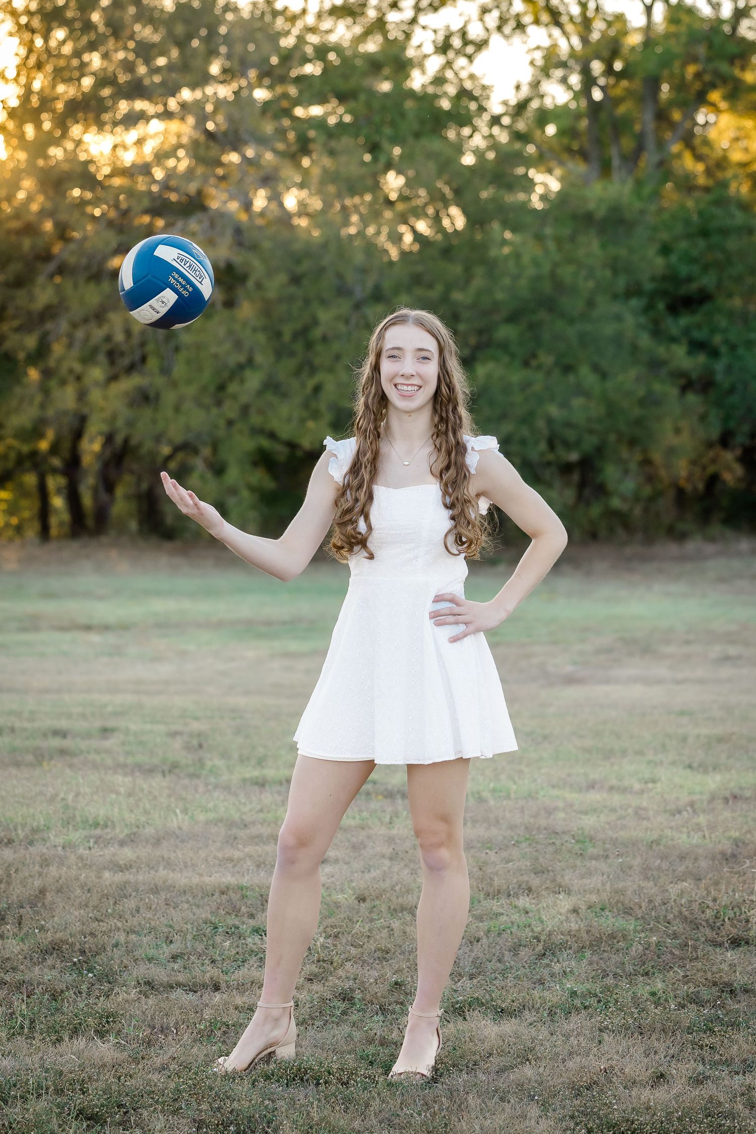 Girl in white dress playing with volleyball on grassy field at sunset.