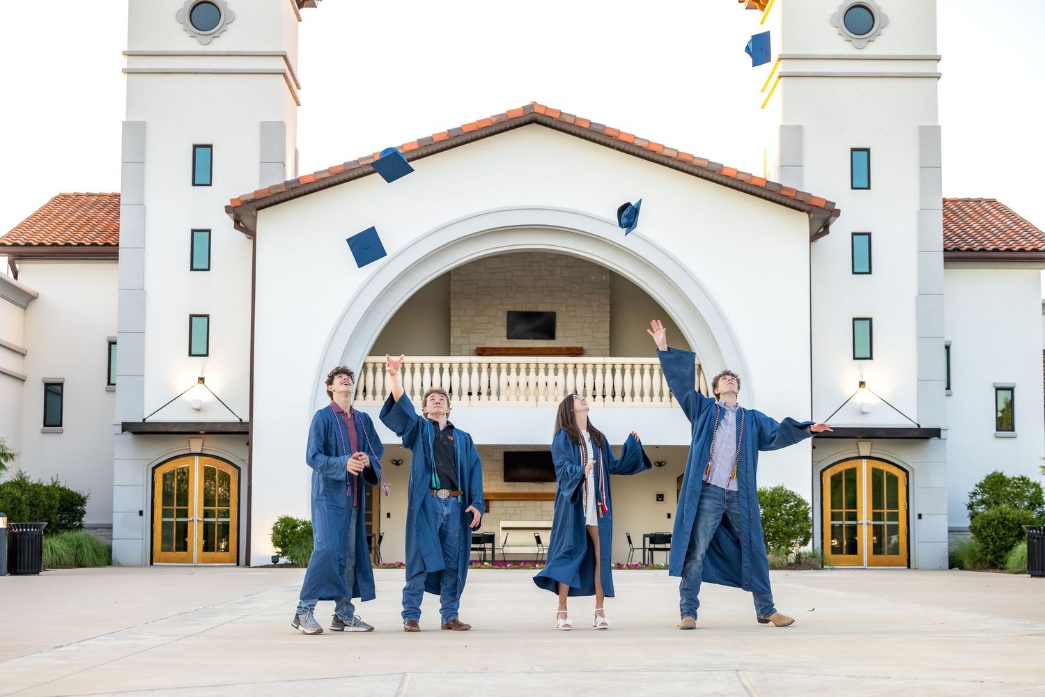Four graduates in blue robes toss caps in front of a white Spanish-style building on graduation day.