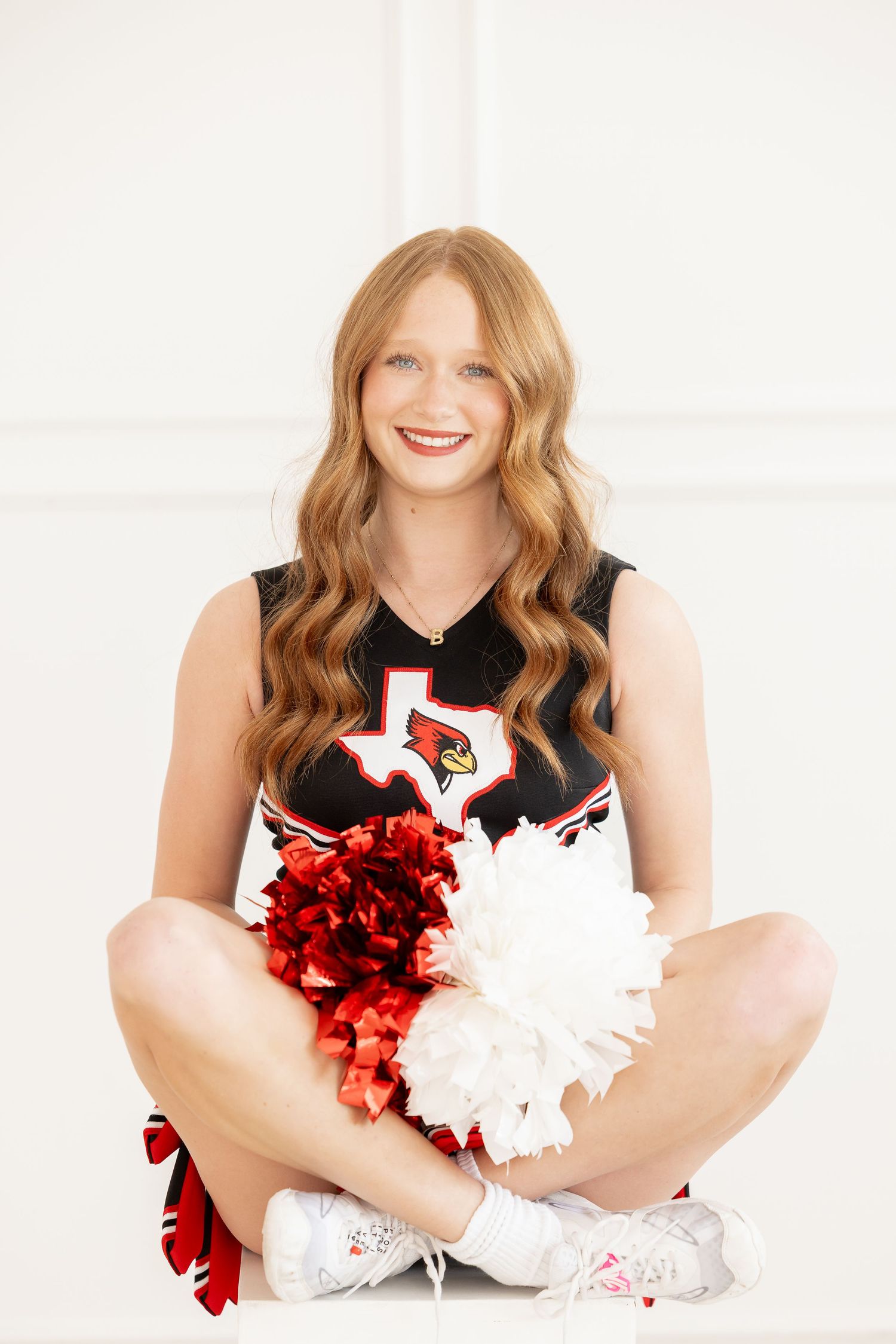 A cheerleader in a red and black uniform sits cross-legged holding red and white pom poms.