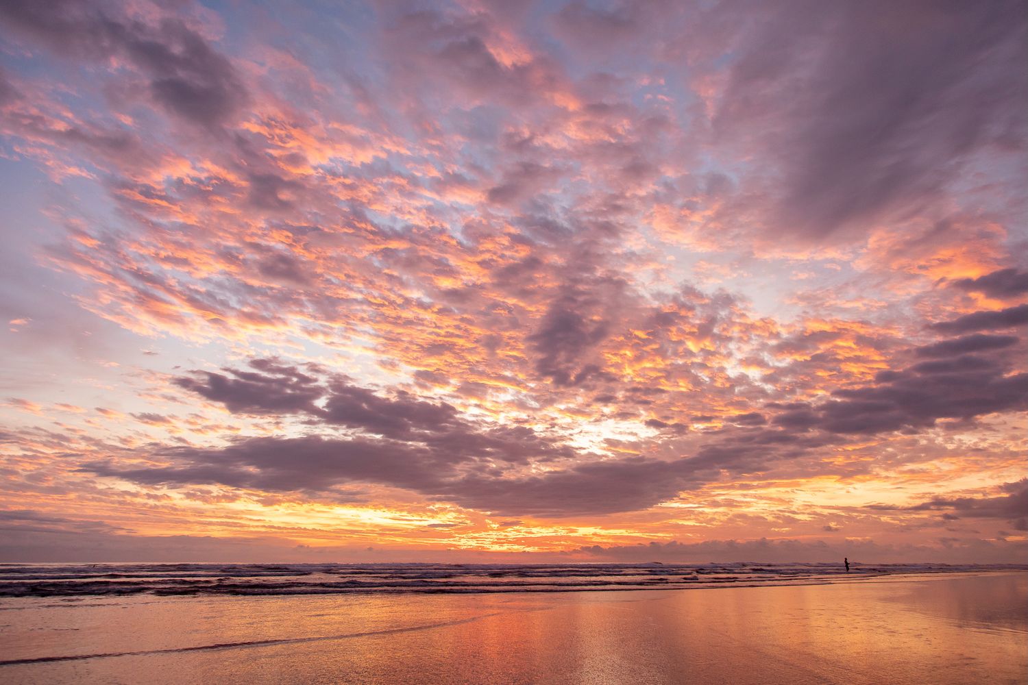 Vibrant orange and pink sunset clouds reflect off wet sand at a peaceful beach during golden hour.