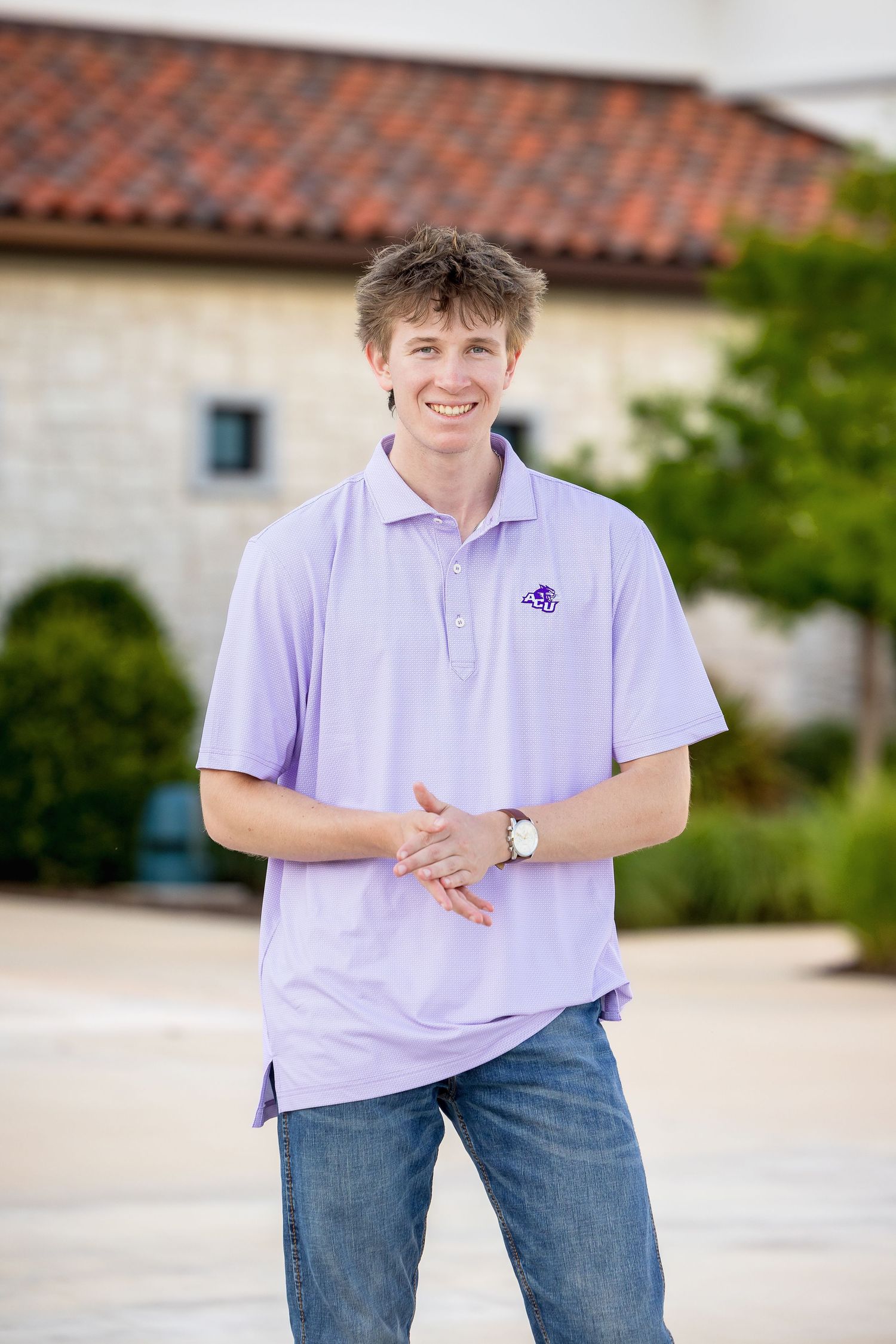 Young man smiling in lavender polo shirt with purple logo, jeans, and watch, posing outdoors near a building.