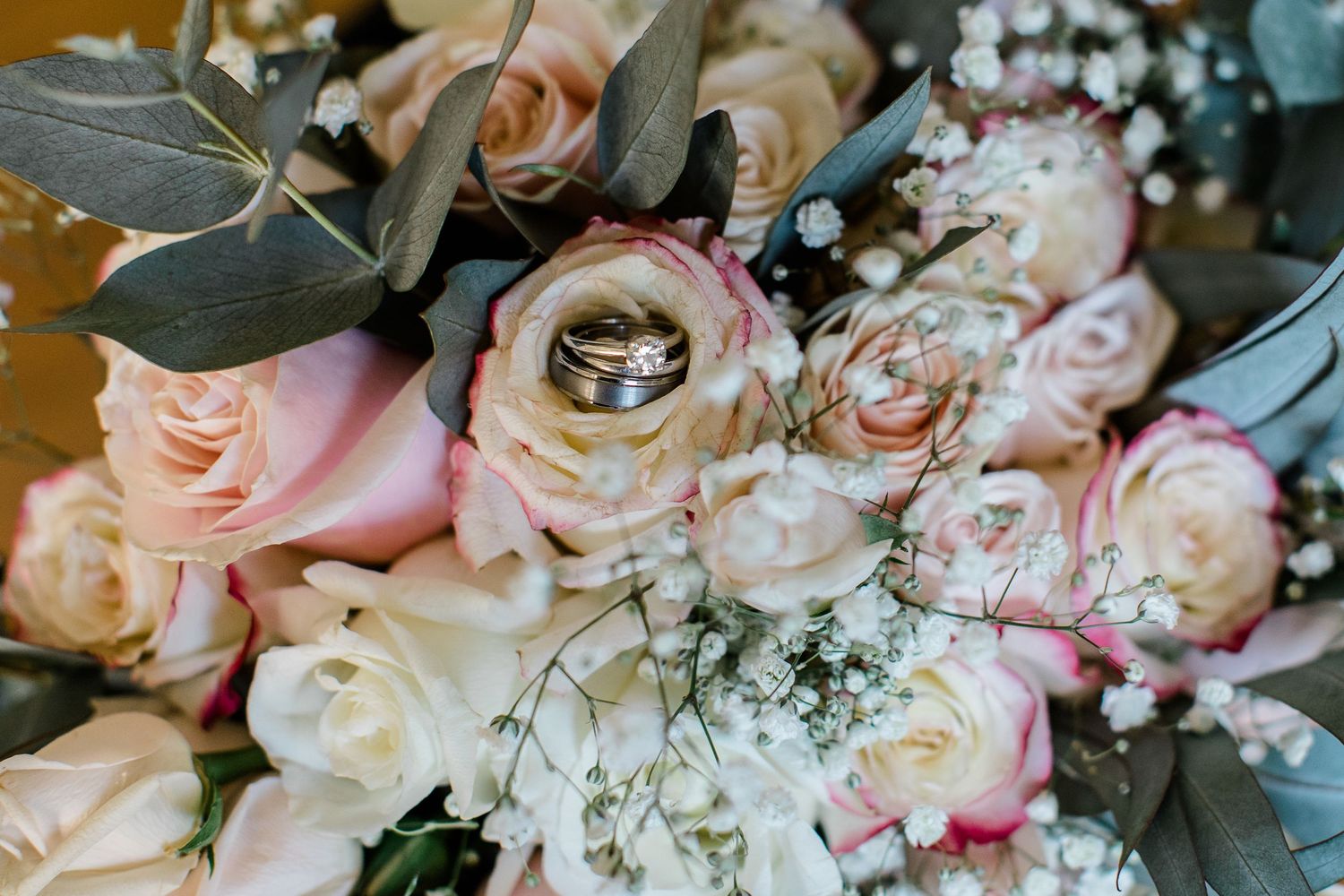 Wedding rings nestled among delicate pink and cream roses with eucalyptus leaves and baby's breath flowers in a bouquet.