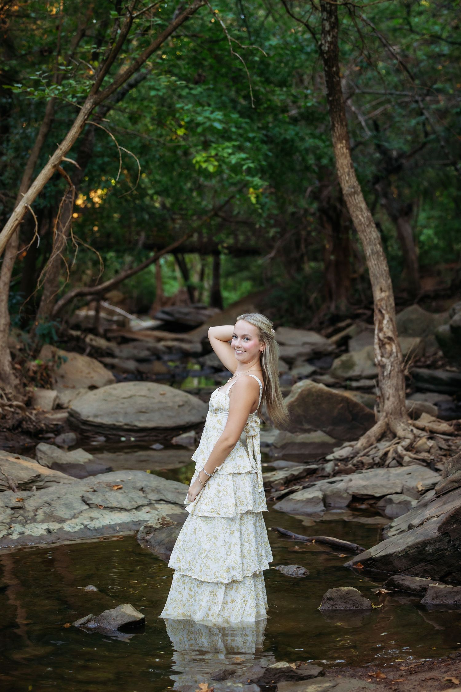 Young girl in white dress wading through creek water surrounded by trees and rocks.