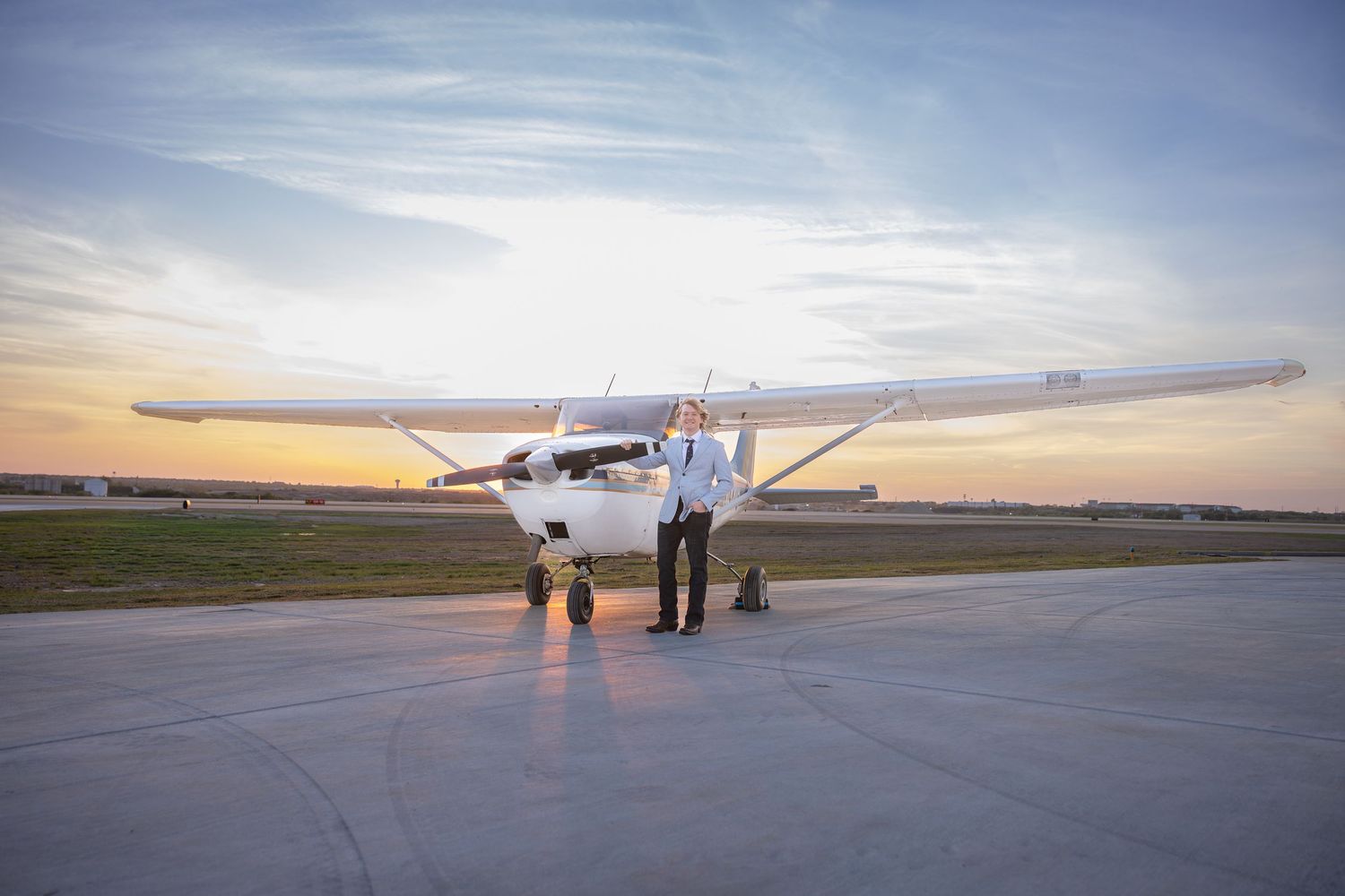 Pilot standing in front of a small Cessna airplane on a runway at sunset with a colorful sky backdrop.