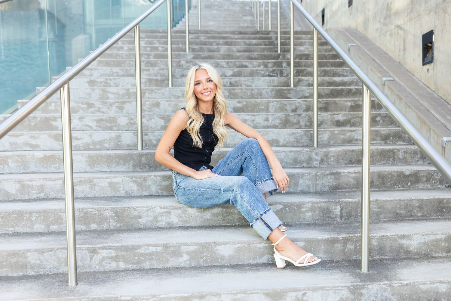 Smiling blonde woman in black top and jeans sitting on concrete stairs with metal railings outdoors.