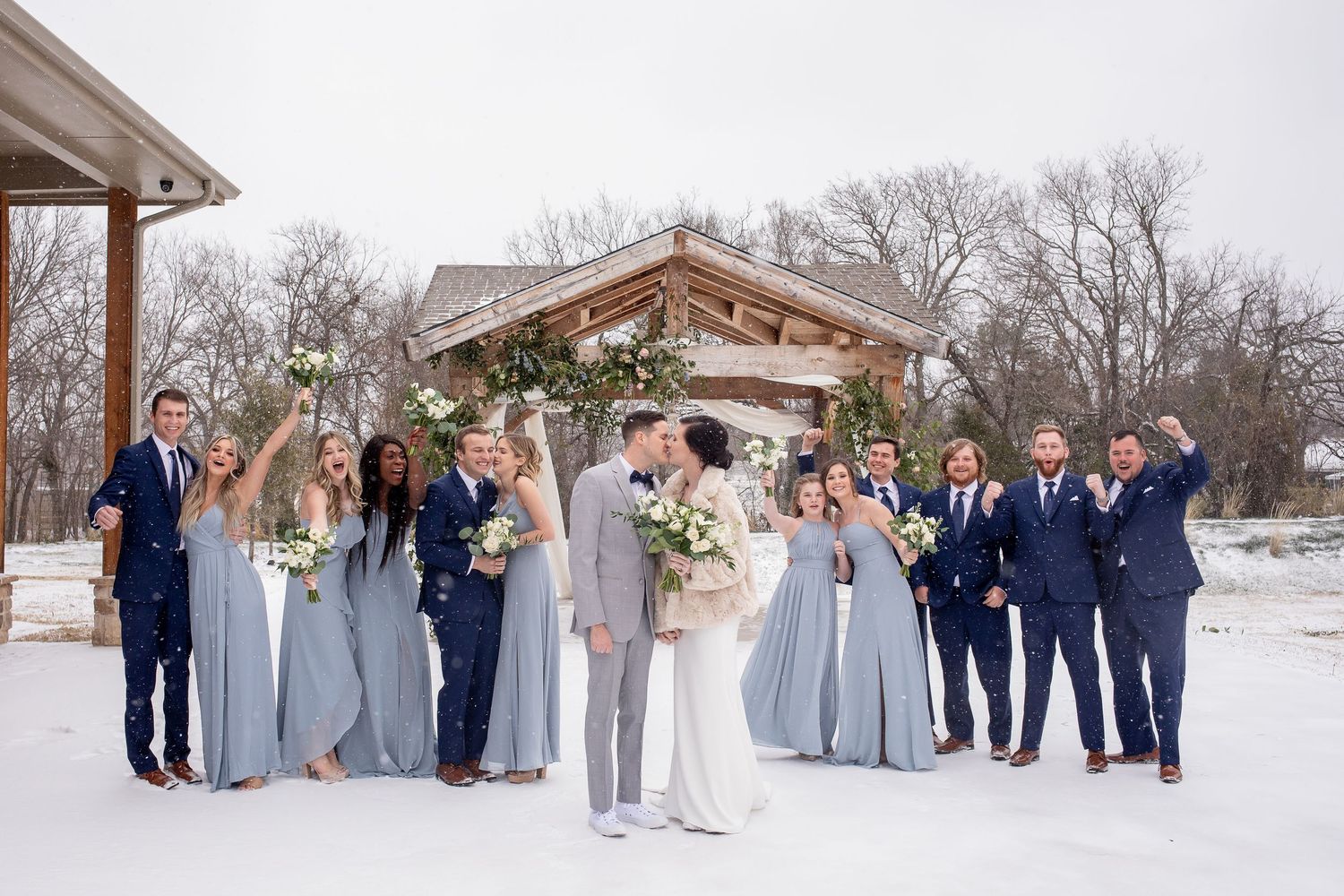 Winter wedding party in blue and gray attire poses together in snow outside rustic wooden gazebo for group photo.