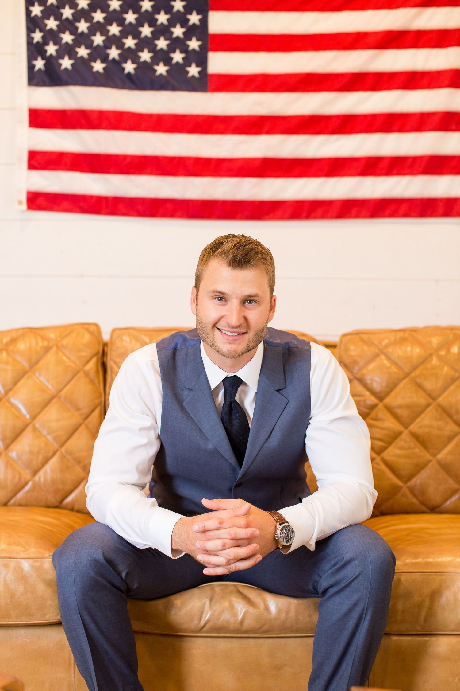 Professional in business attire sits on brown leather couch with American flag backdrop in office setting.