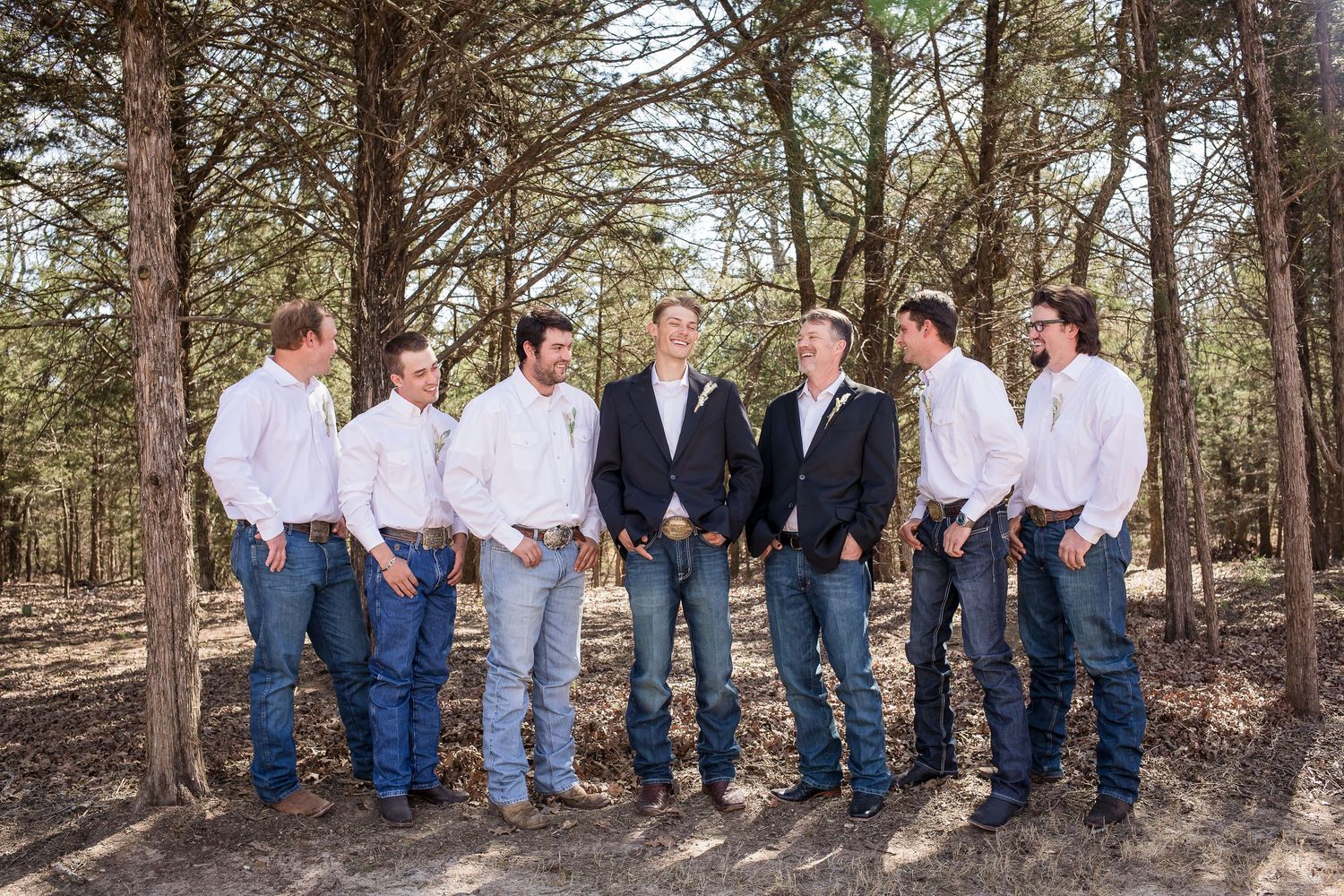 A group stands together in a wooded setting wearing coordinated outfits of white shirts, jeans and navy blazers.
