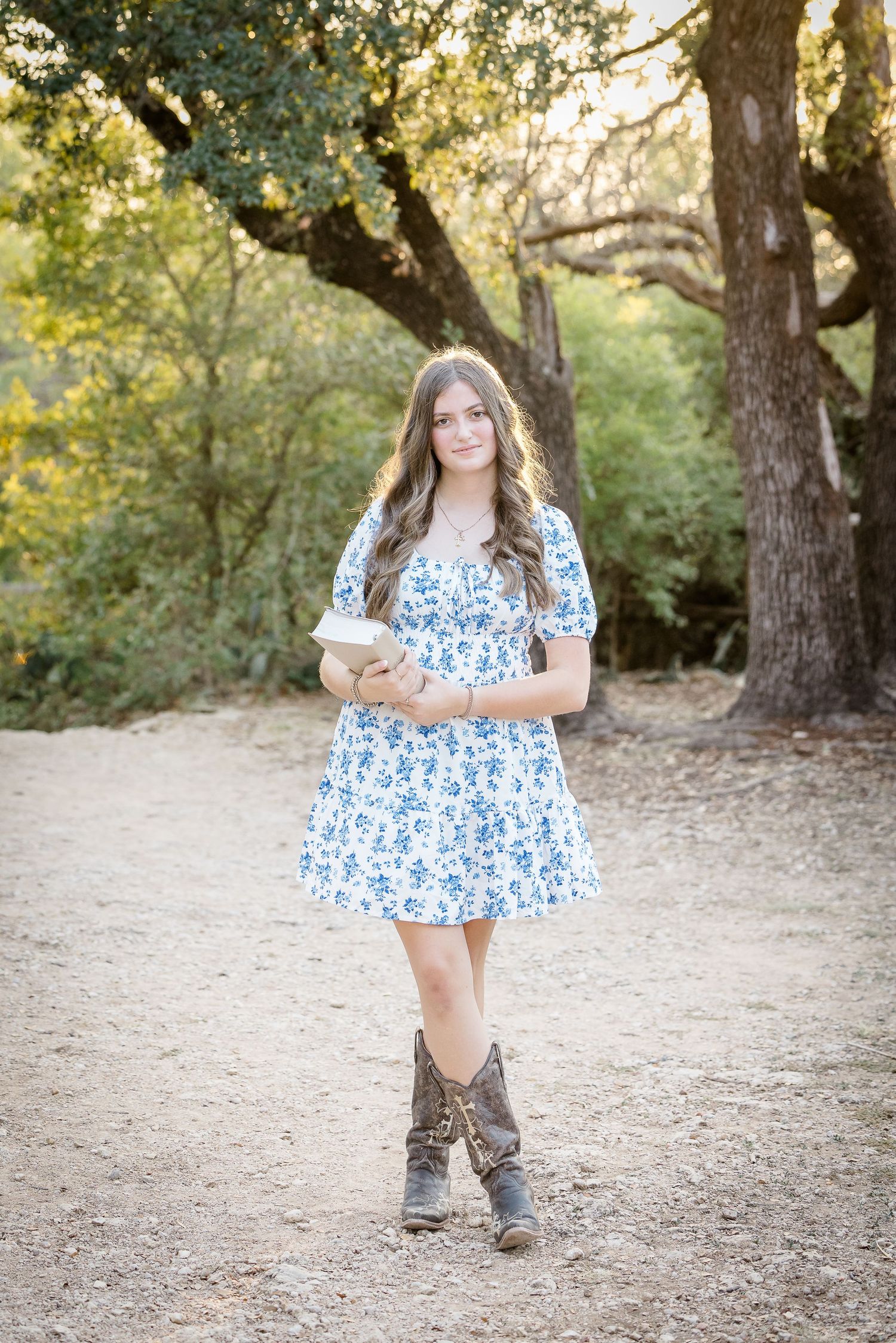 A person in a blue floral dress and boots poses playfully in a wooded outdoor setting.