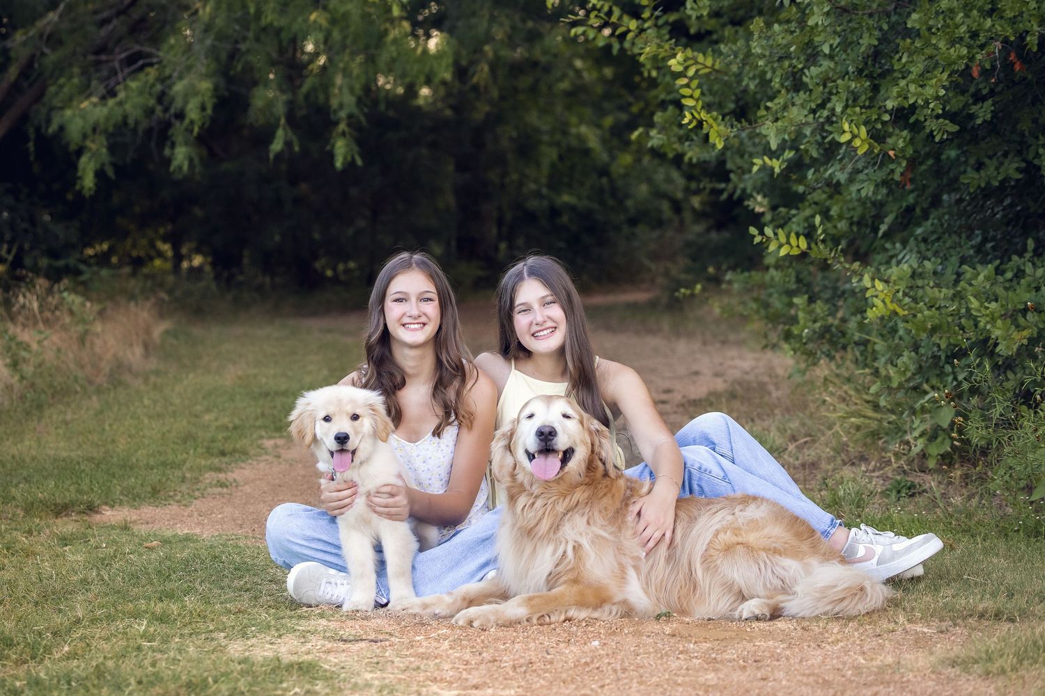 Two people and their golden retrievers pose together on a dirt path in nature.