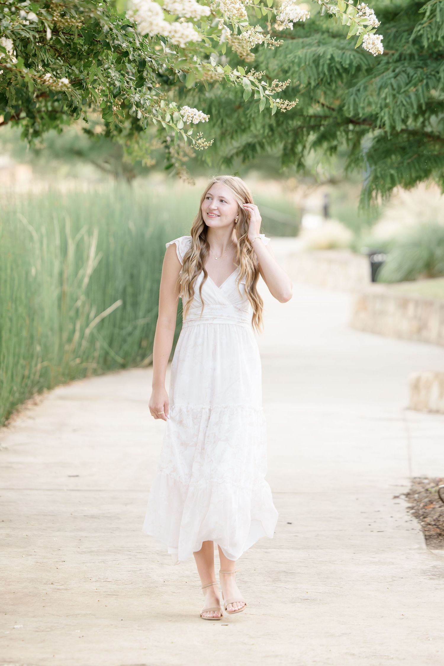 Young woman in white lace dress walking on garden path surrounded by lush greenery and blooming flowers.