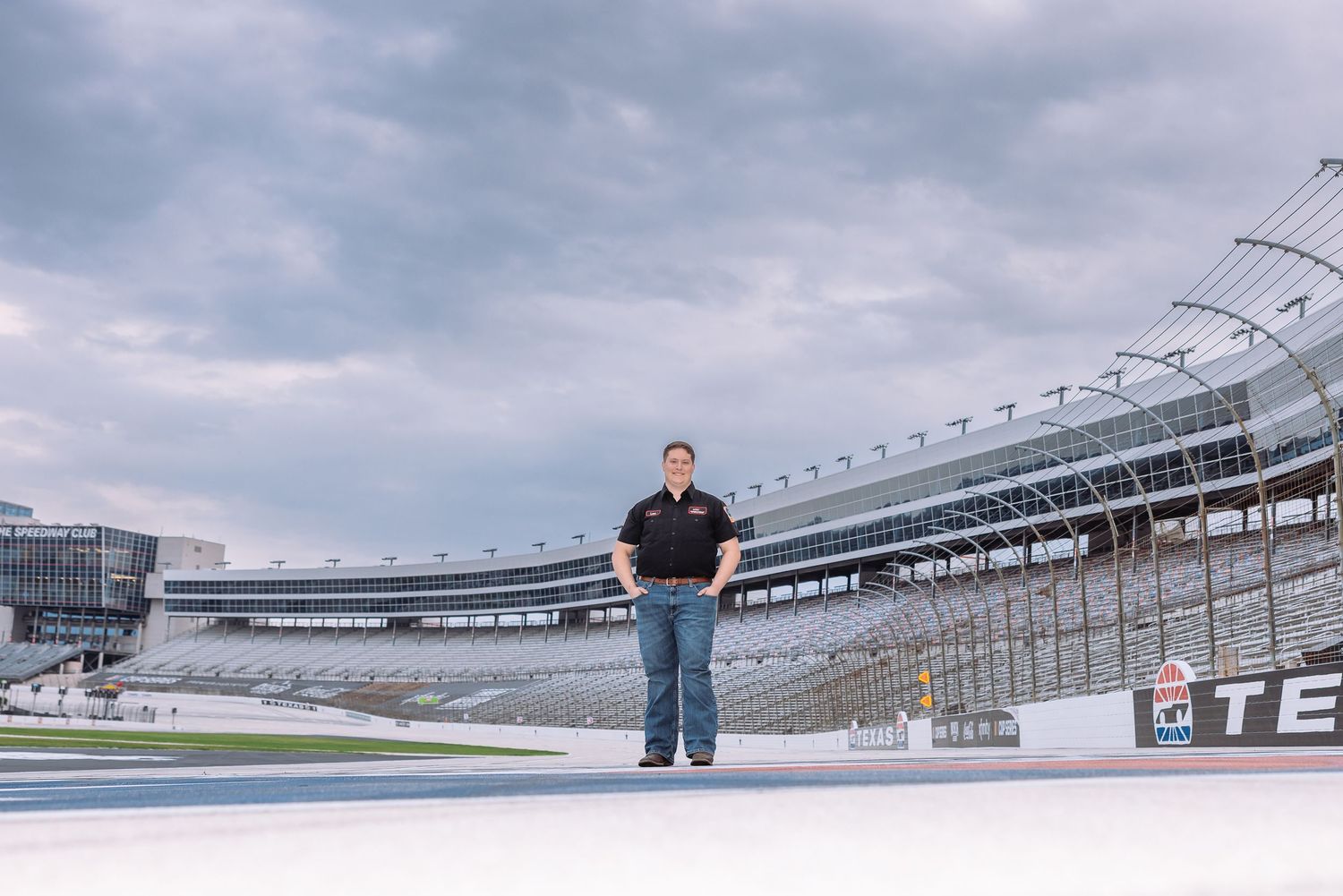 Person standing on NASCAR racetrack infield with empty grandstands and cloudy sky at Texas Motor Speedway.