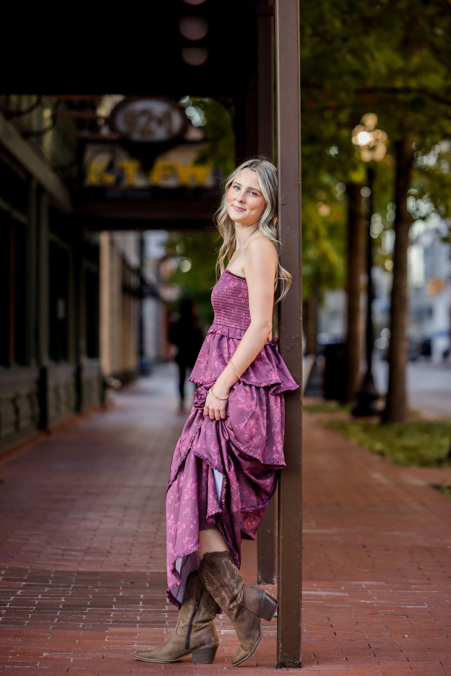 Young woman in purple strapless dress and cowboy boots leaning against a pole on a brick sidewalk downtown.