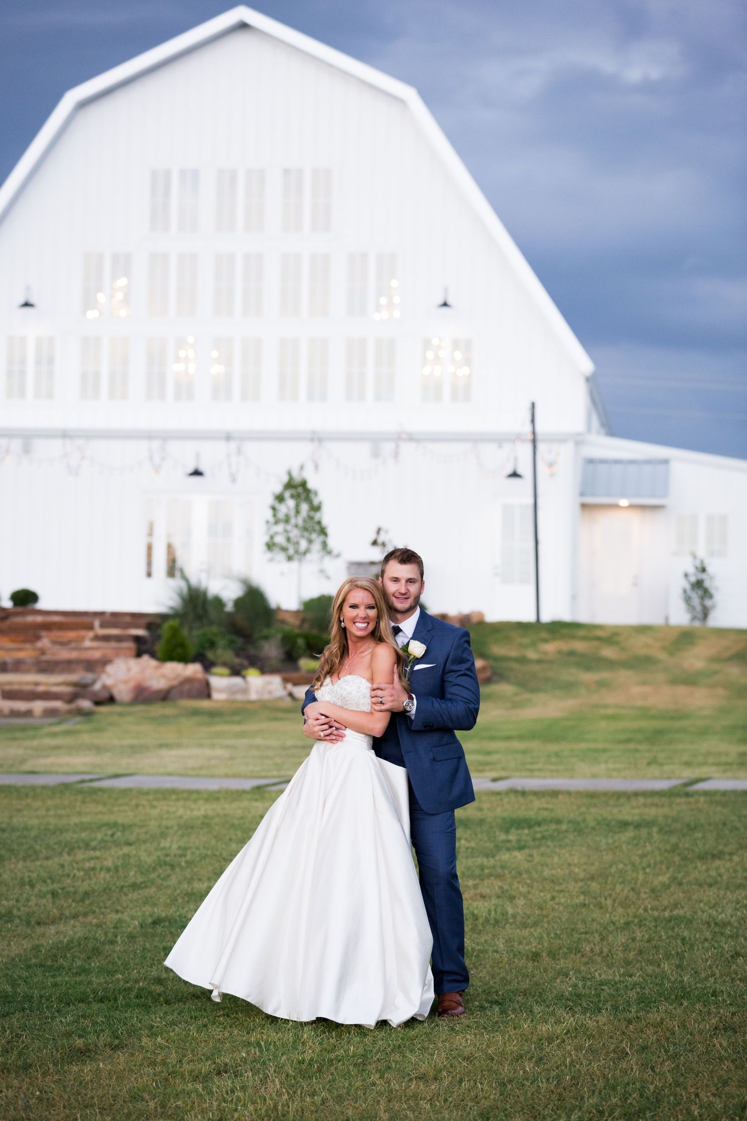 A couple poses for a wedding photo in front of a beautiful white barn wedding venue on a grassy lawn.