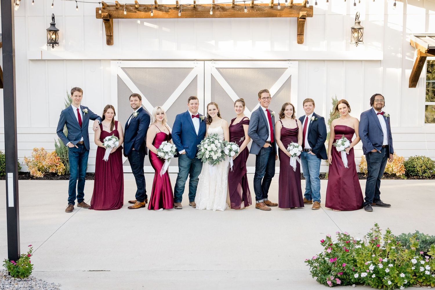 Wedding party in navy suits and burgundy gowns pose together outside a rustic white barn venue.
