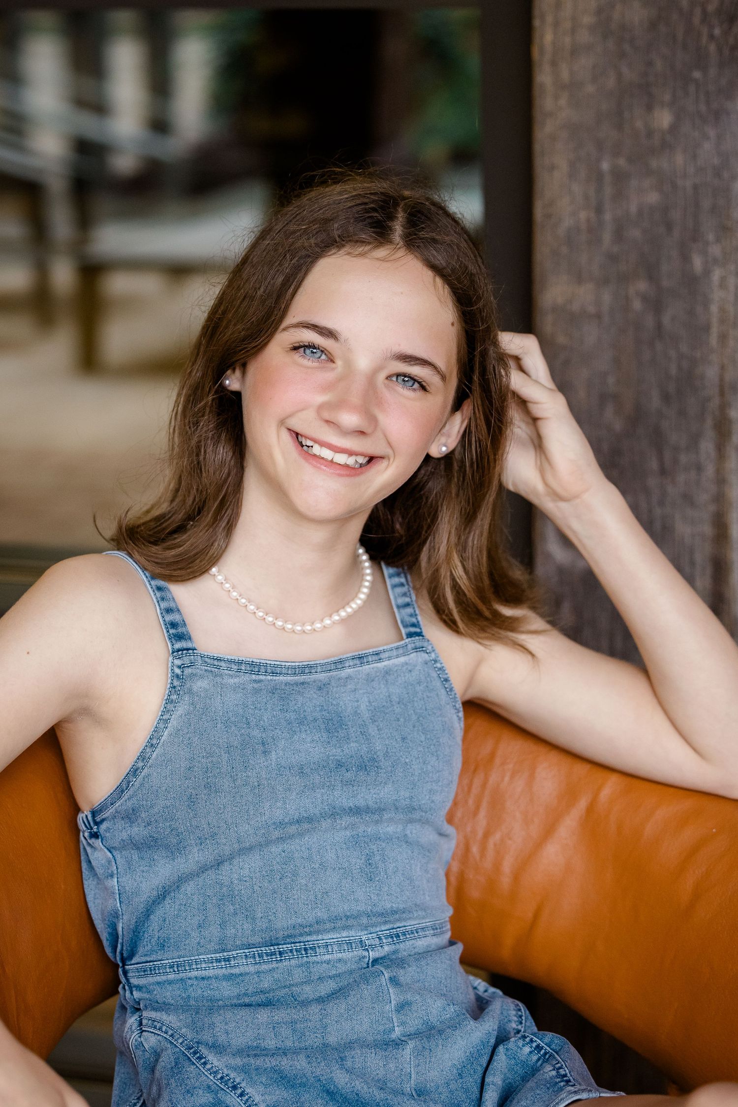 Portrait of someone wearing a denim overall dress and pearl necklace while sitting on a brown leather couch.