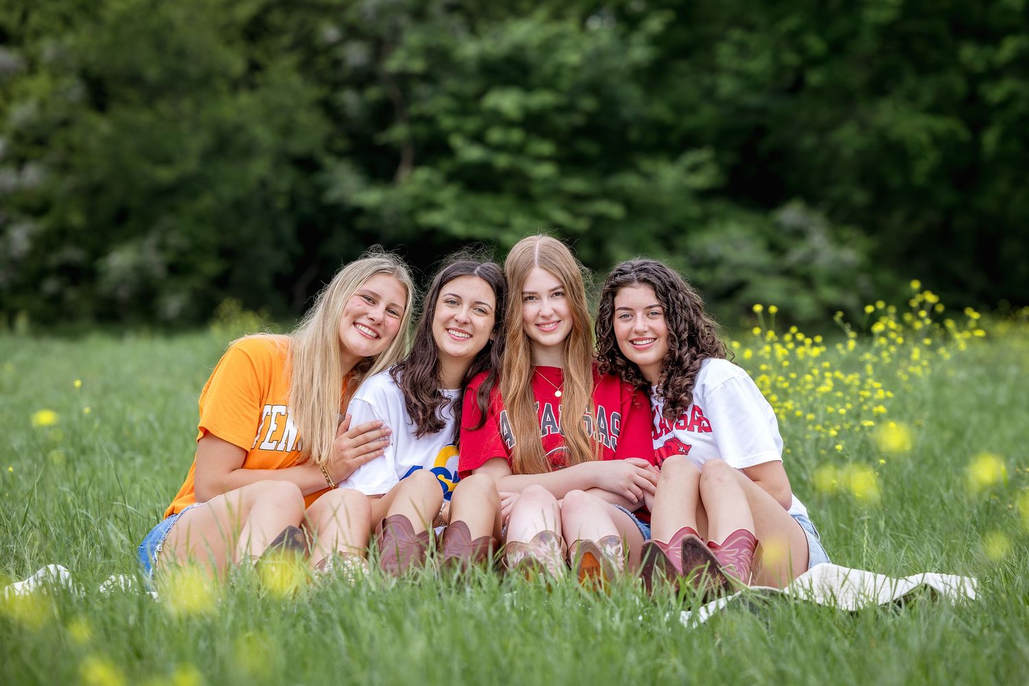 Four smiling young women sitting together in a green meadow with yellow wildflowers and trees in the background.