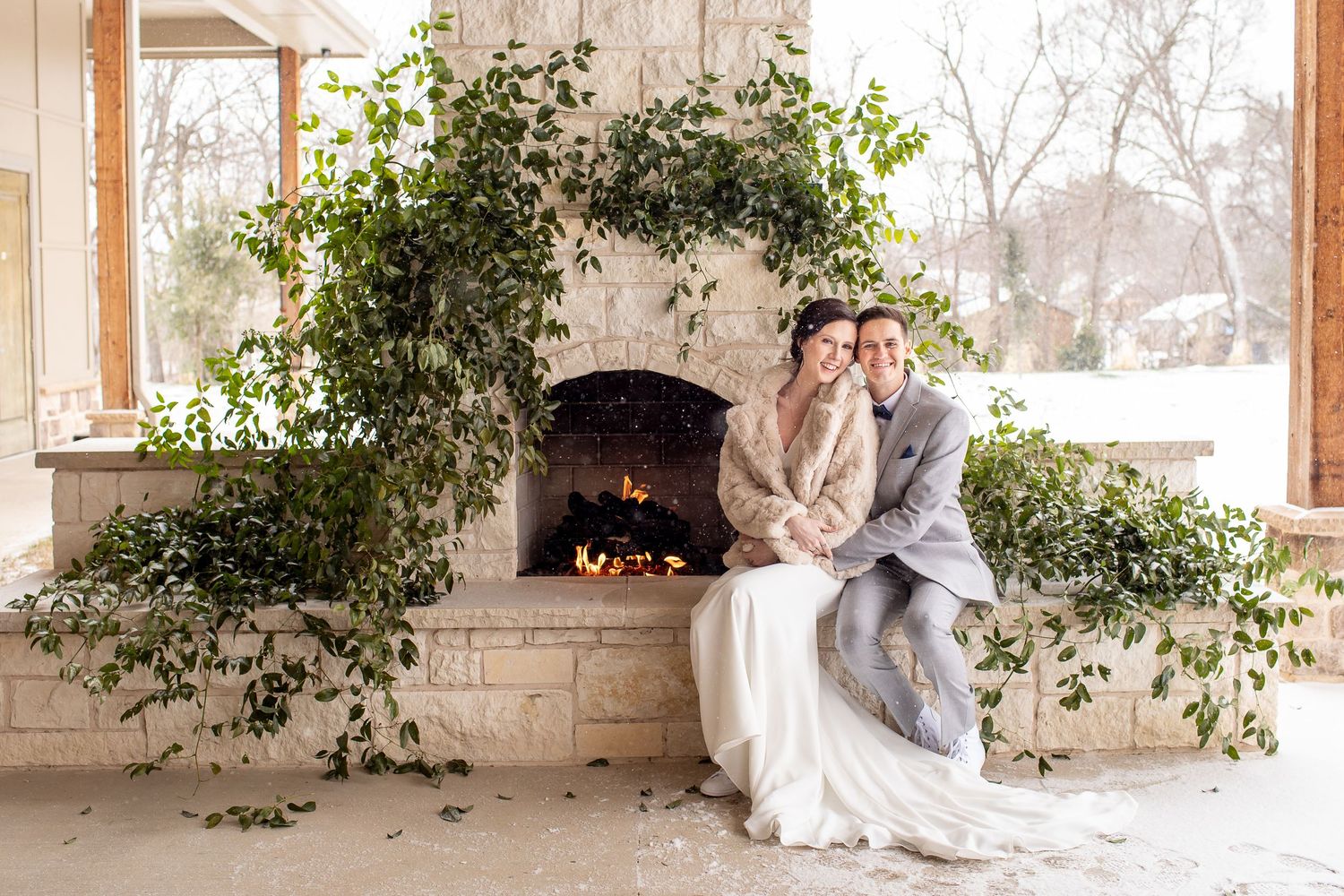 A couple in wedding attire sits intimately by a stone fireplace adorned with cascading greenery in an elegant outdoor setting.