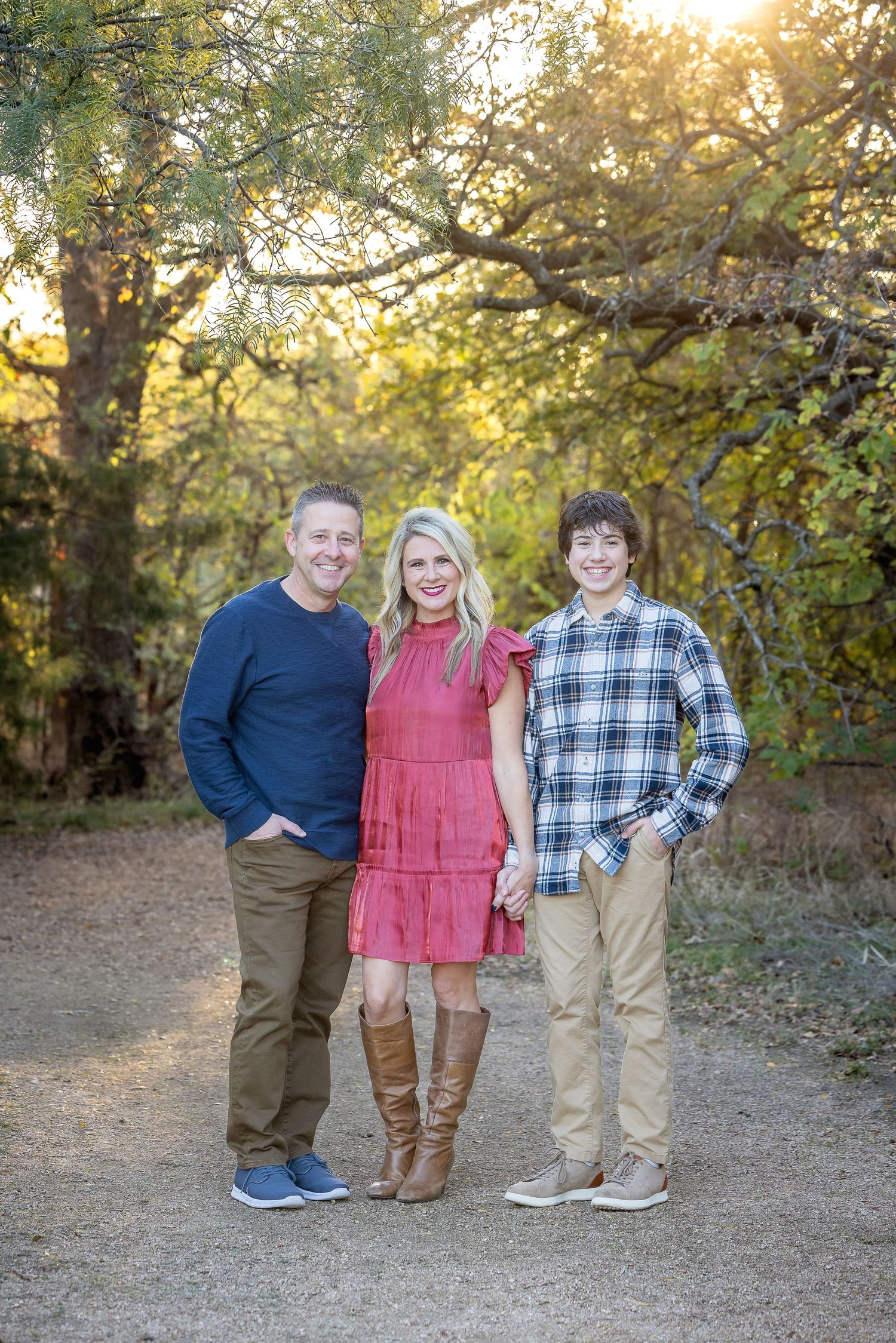 Family portrait of three people standing on a dirt path surrounded by autumn trees at sunset.