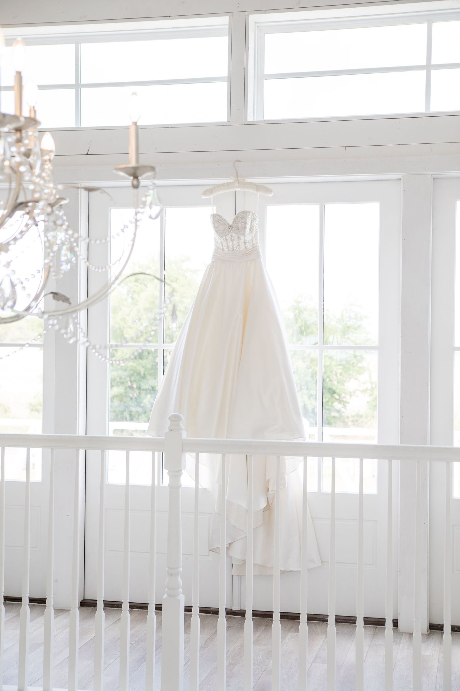 An elegant white wedding dress hangs in a bright sunlit room with crystal chandeliers and white railings.