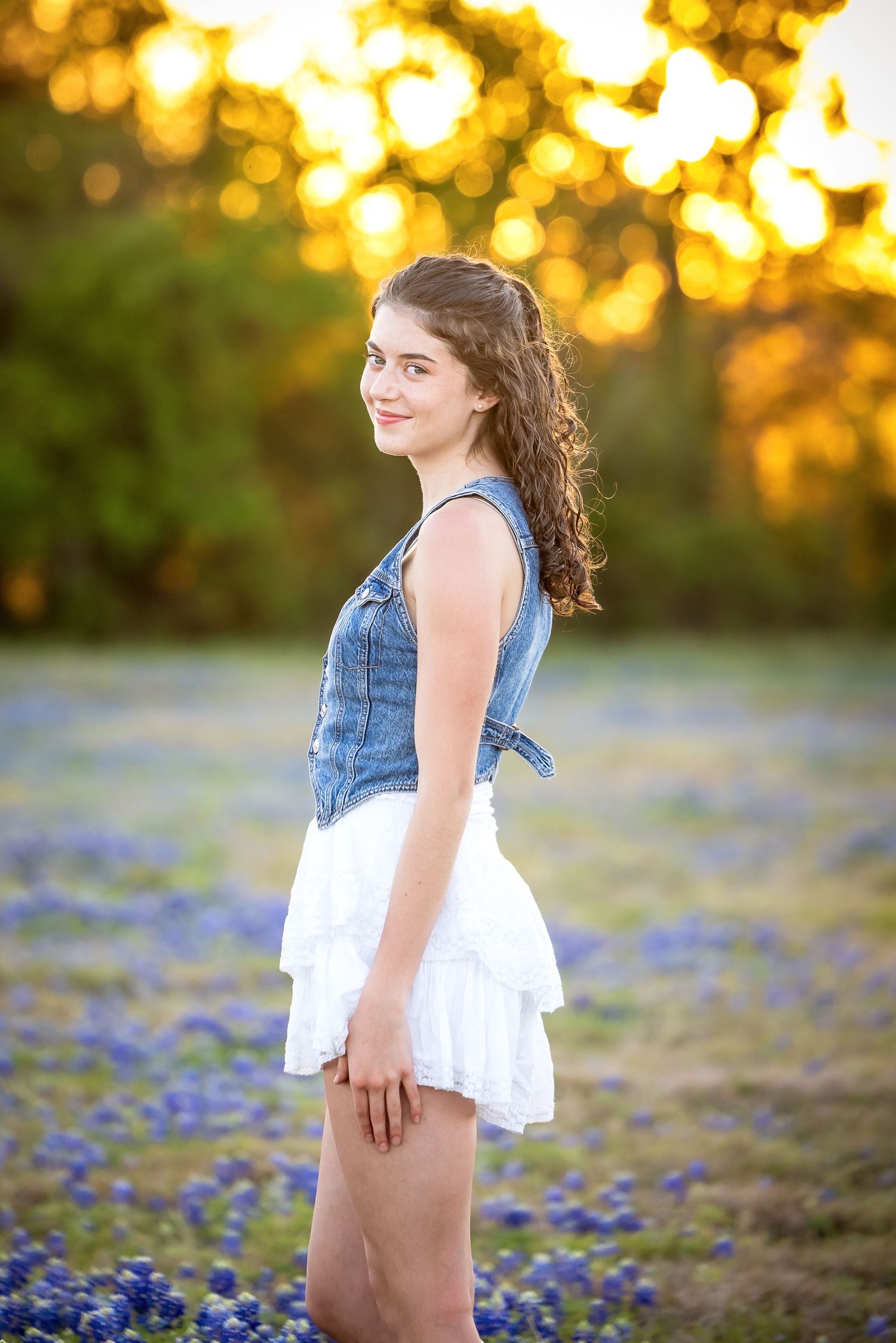 Young woman in denim vest and white skirt smiling in a field of Texas bluebonnets at golden hour.