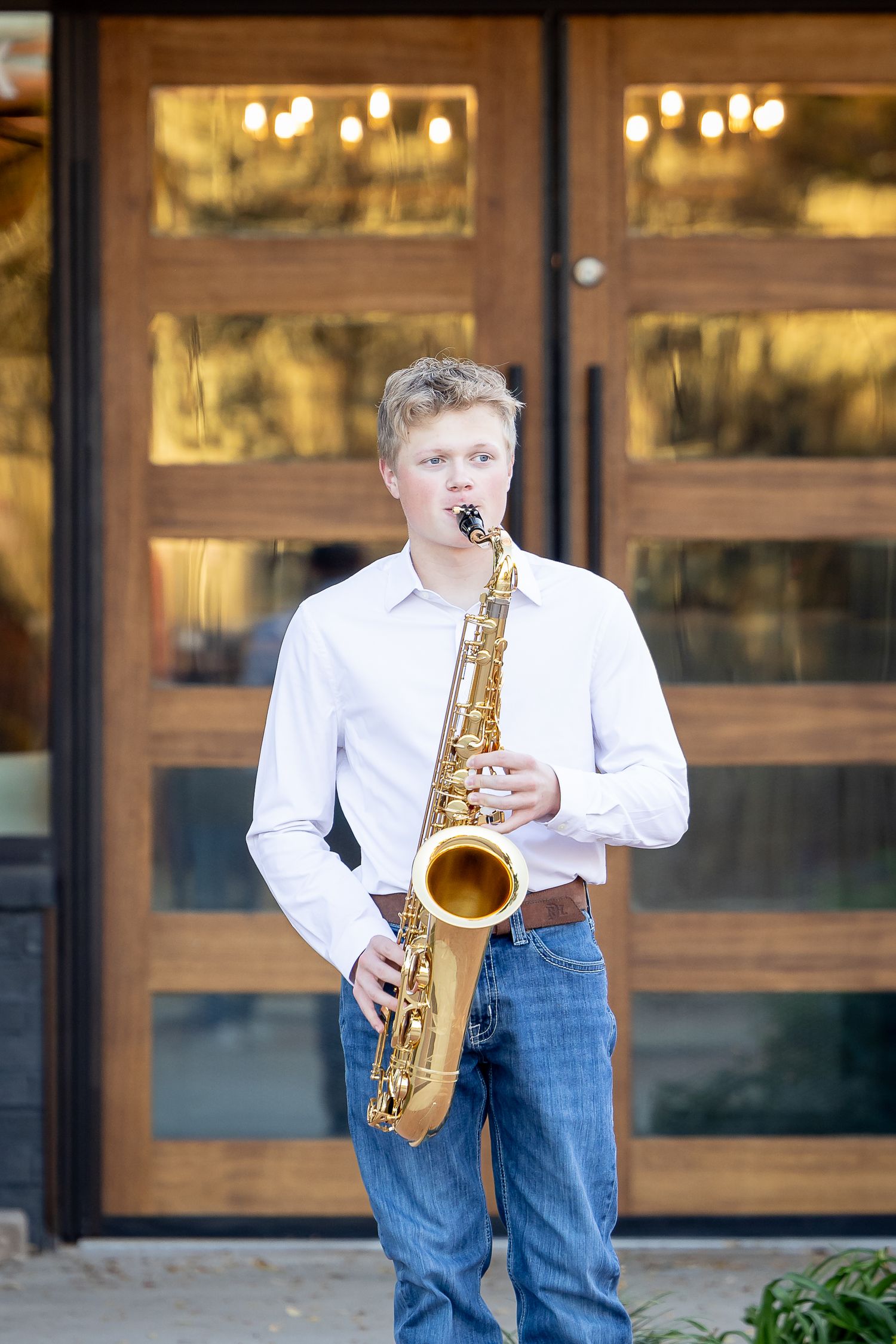 A young musician in white shirt and jeans plays saxophone against a building exterior.